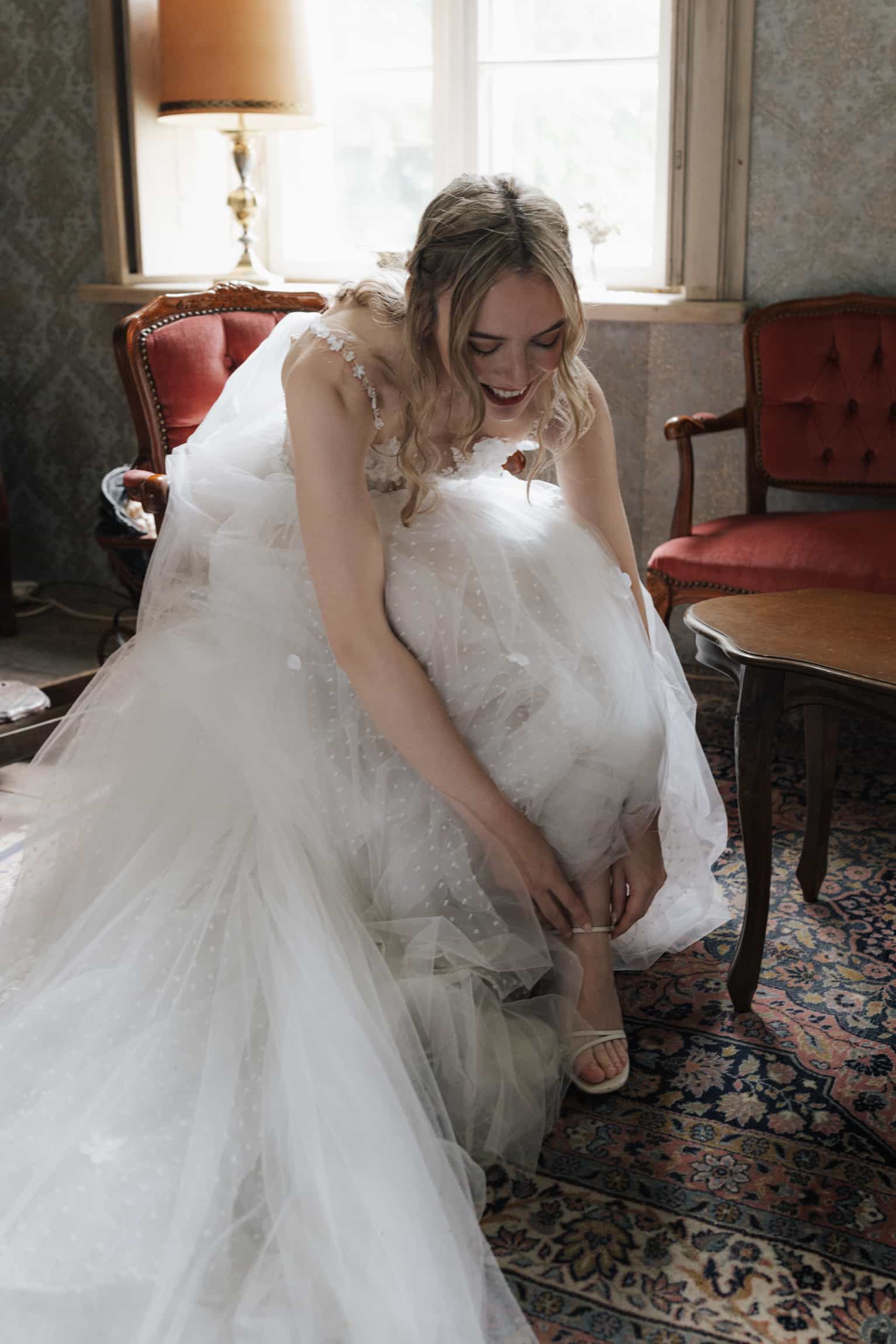 Braut im weißen Spitzenkleid richtet ihr Kleid in einem eleganten Hotelzimmer mit Vintage-Möbeln