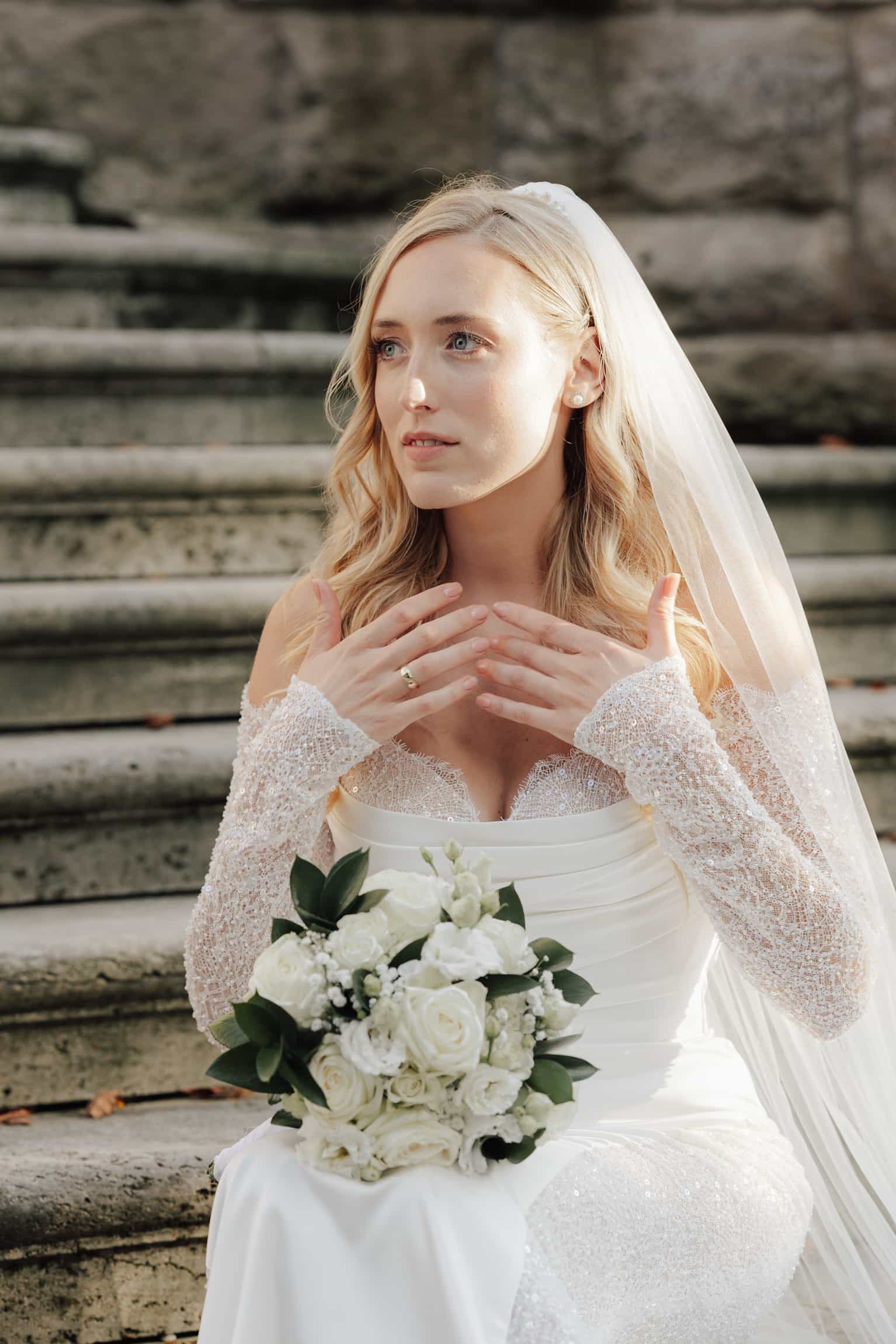 Blonde Braut im weißen perlenbesetzten Kleid und Schleier sitzt nachdenklich auf verwitterten Steintreppen