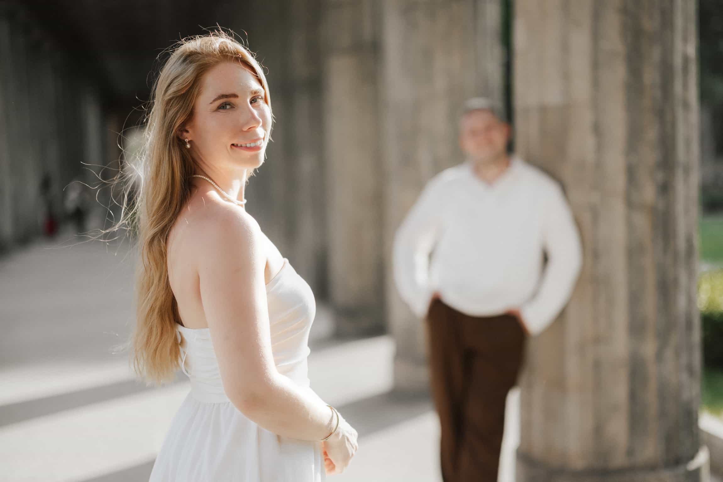 Blonde Braut im weißen Kleid lächelt im warmen Sonnenlicht, Bräutigam unscharf vor Steinsäulen dahinter