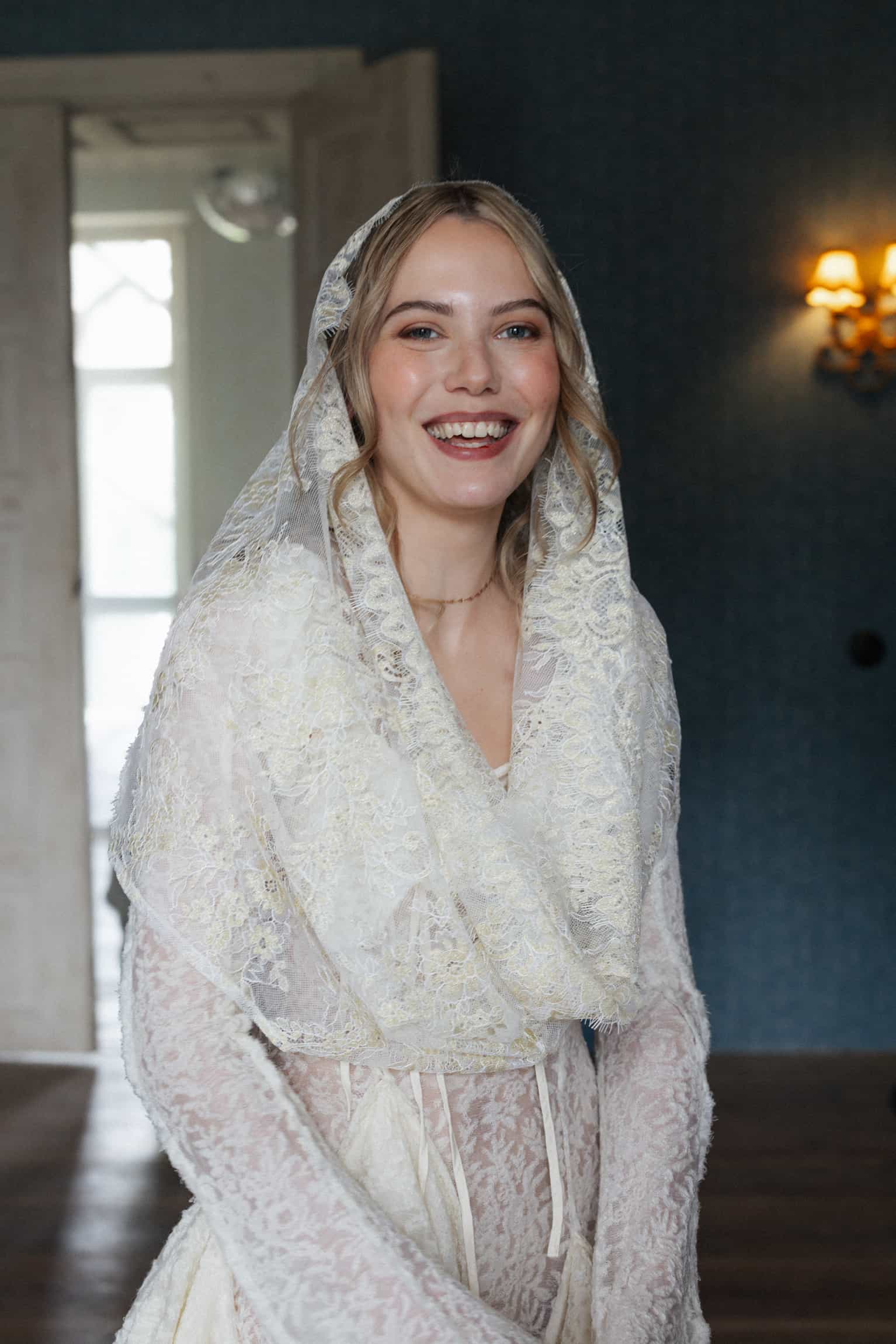 Braut mit zartem Spitzenschleier und Spitzenkleid, warm lächelnd im Innenraum bei goldenem Licht