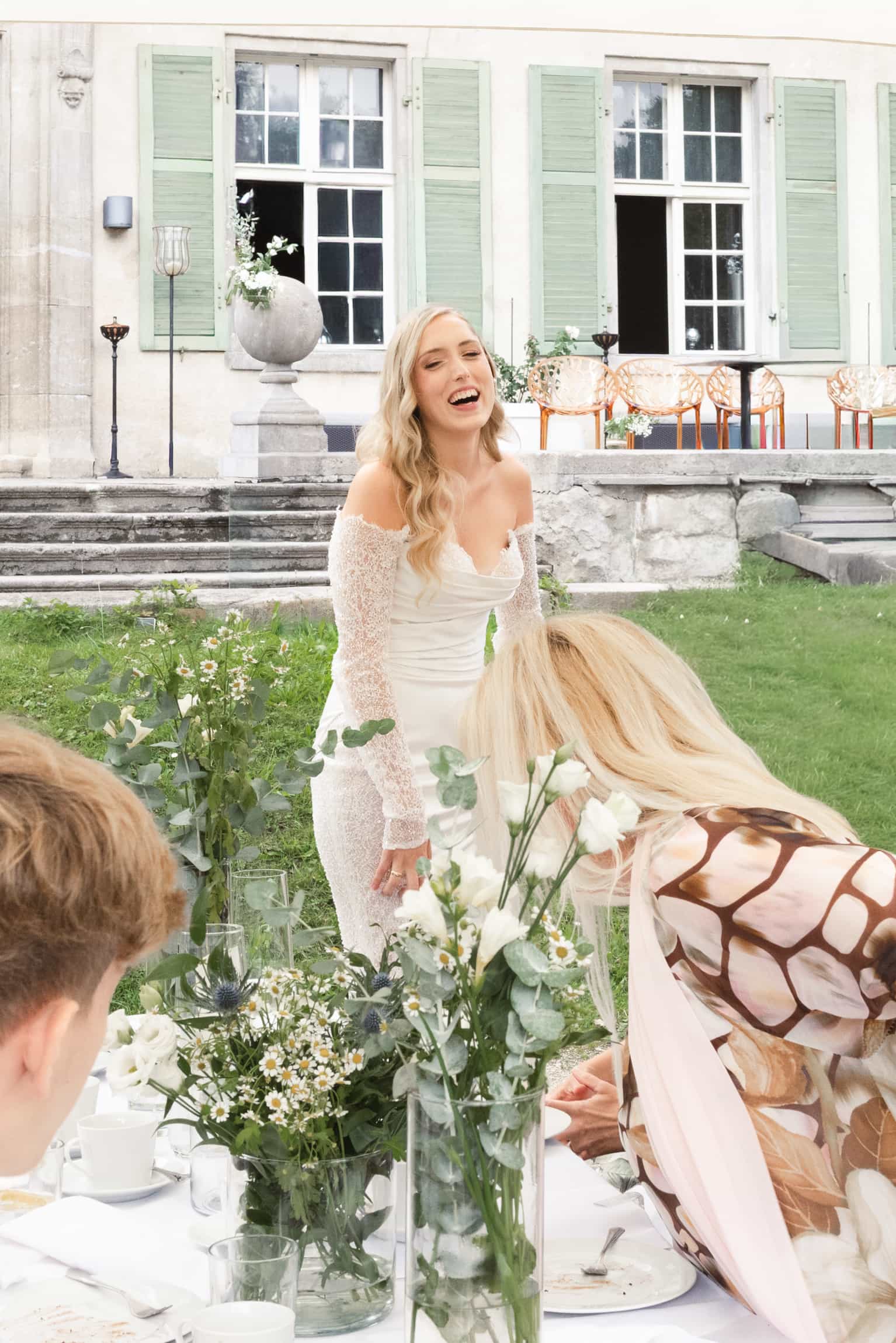 Braut im weißen Hochzeitskleid lacht bei einer Gartenfeier vor einem klassischen Haus