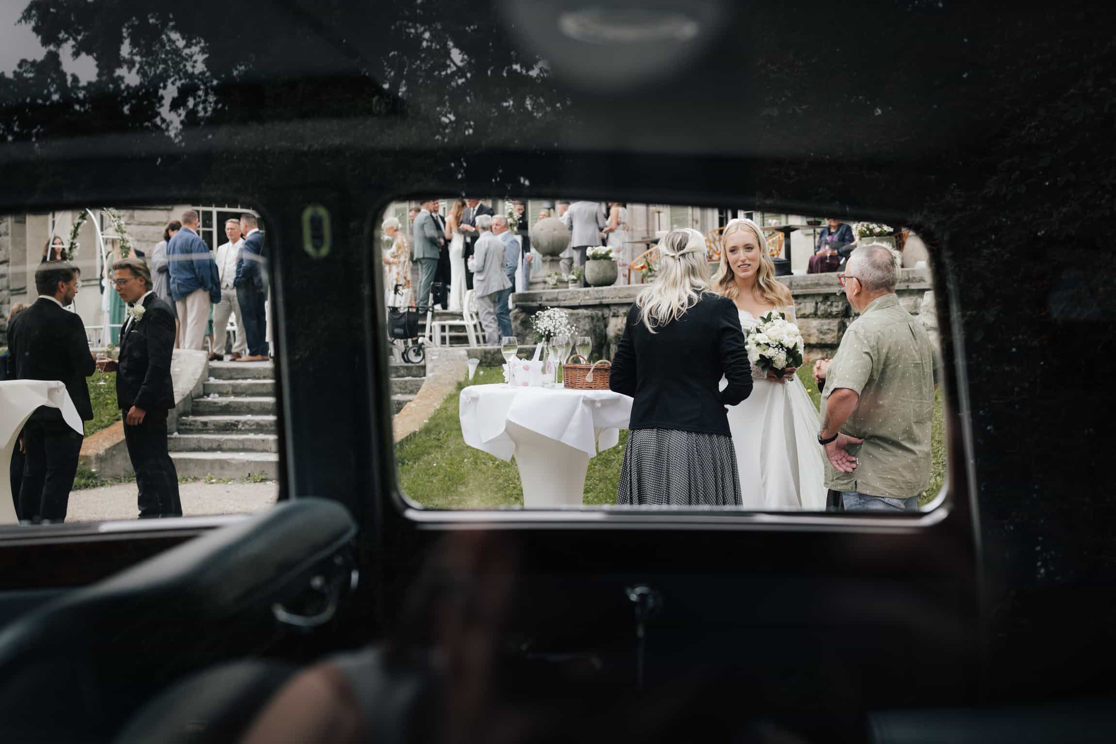 Hochzeitsgäste und Feier durch ein Autofenster, Frau mit blondem Haar und Blumen im Vordergrund
