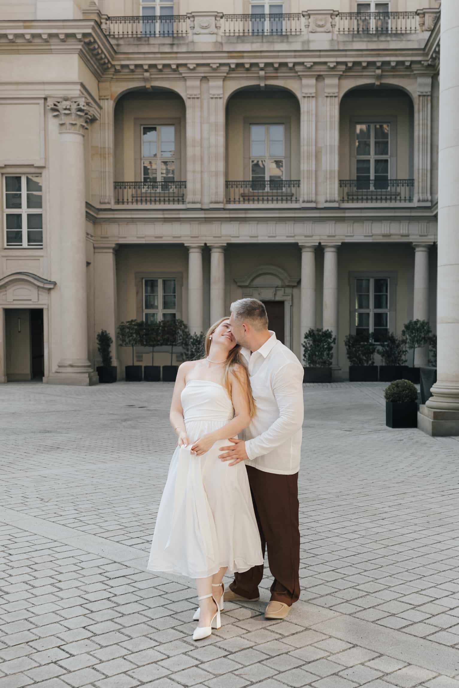 Bräutigam im weißen Hemd küsst lächelnde Braut im weißen Kleid in einem weiten klassizistischen Schlosshof