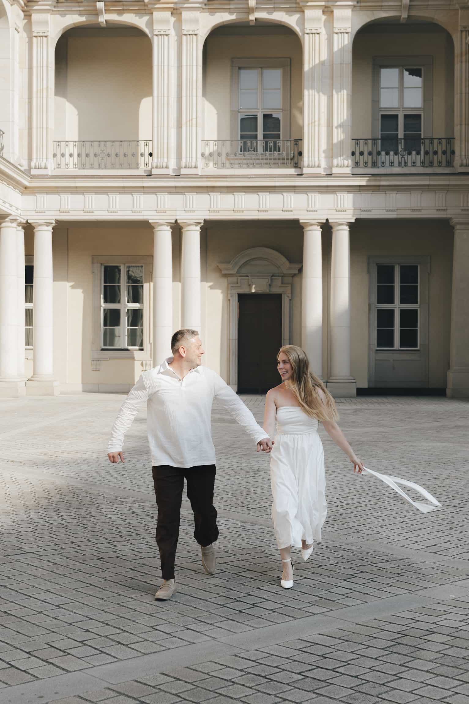 Braut im weißen Kleid und Bräutigam laufen Hand in Hand über einen gepflasterten Hof vor klassischer Schlossfassade