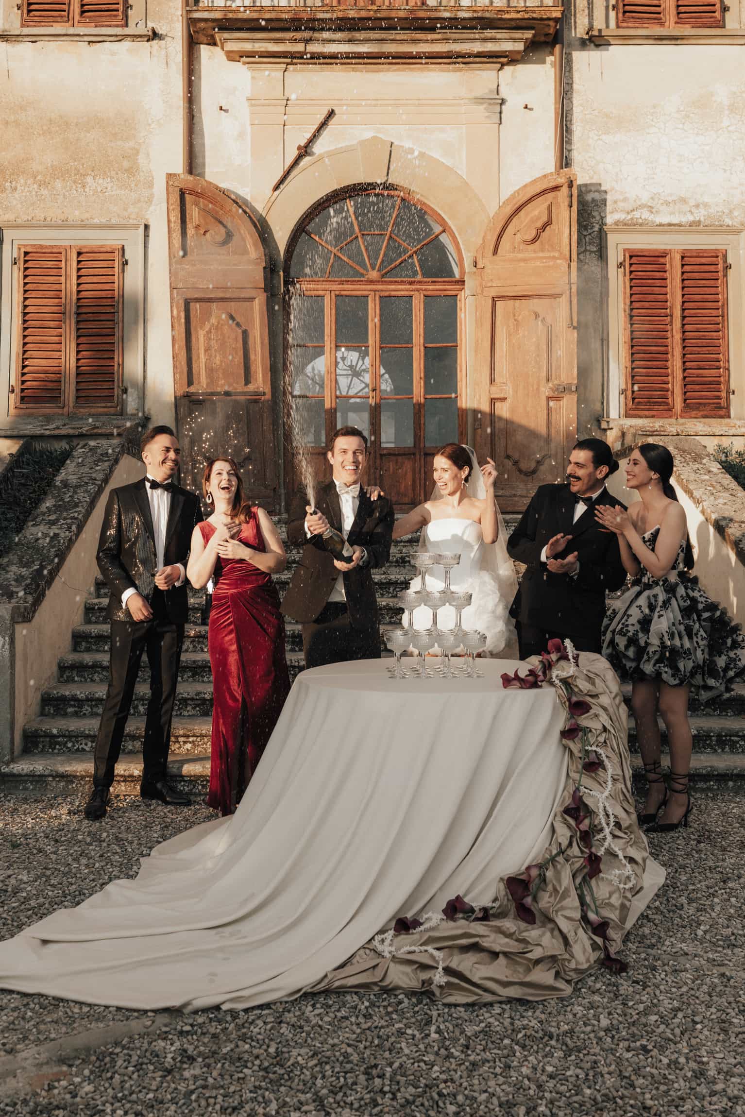 Braut mit langer Schleppe in weißem Kleid vor einer klassischen Villa mit Gästen
