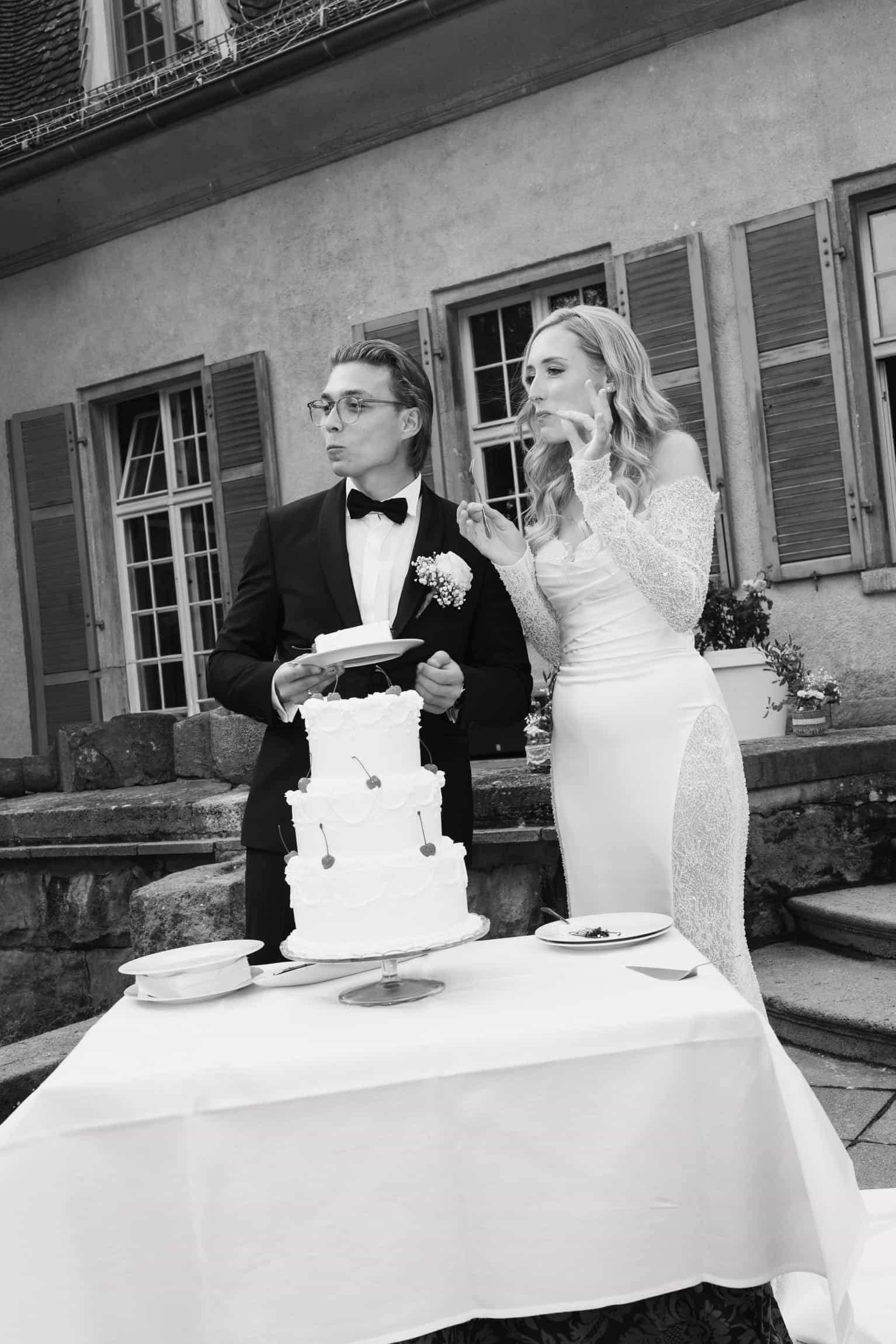Braut berührt Bräutigams Gesicht, während er die Hochzeitstorte hält, klassische Architektur im Hintergrund