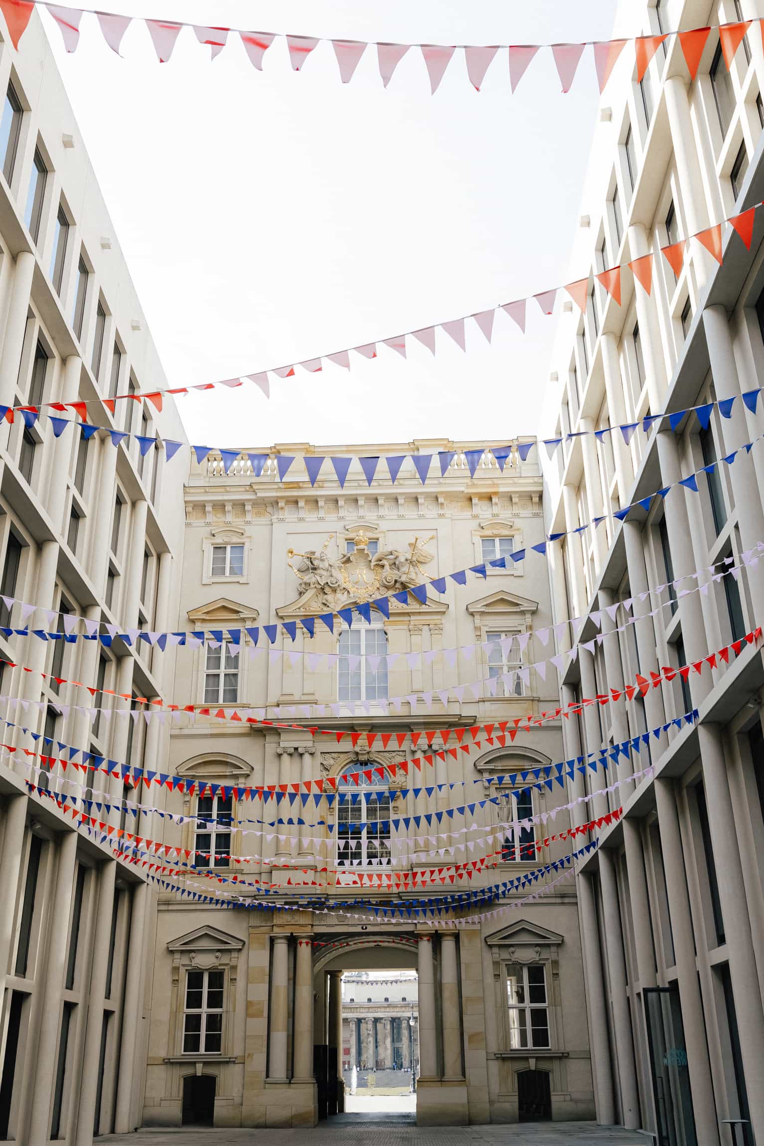 Bunte dreieckige Wimpelketten über einem Innenhof, der zu einem barocken Schlossportal mit goldenem Wappen führt