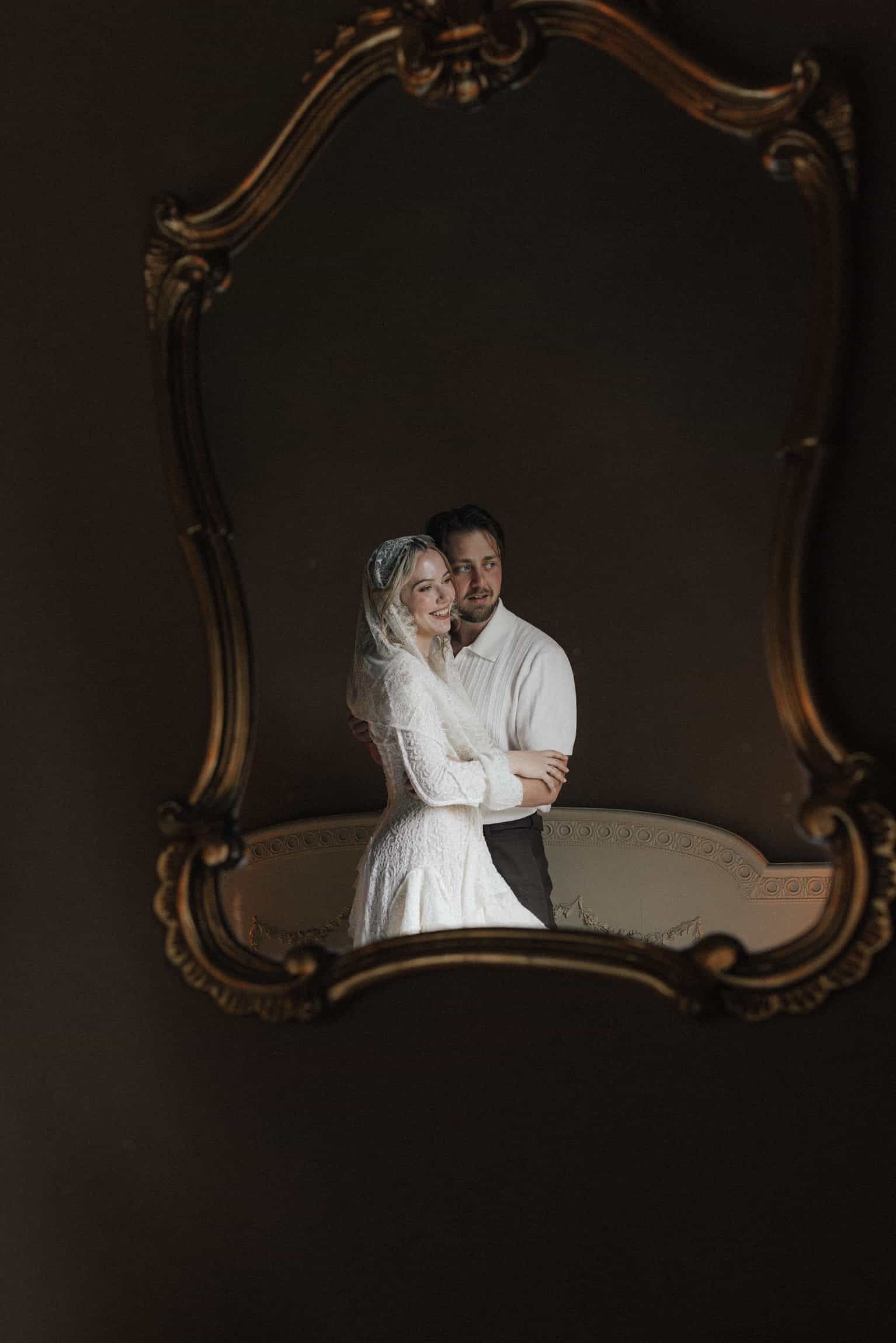 Bride in lace veil and groom framed by an ornate gilt baroque mirror against a dark backdrop