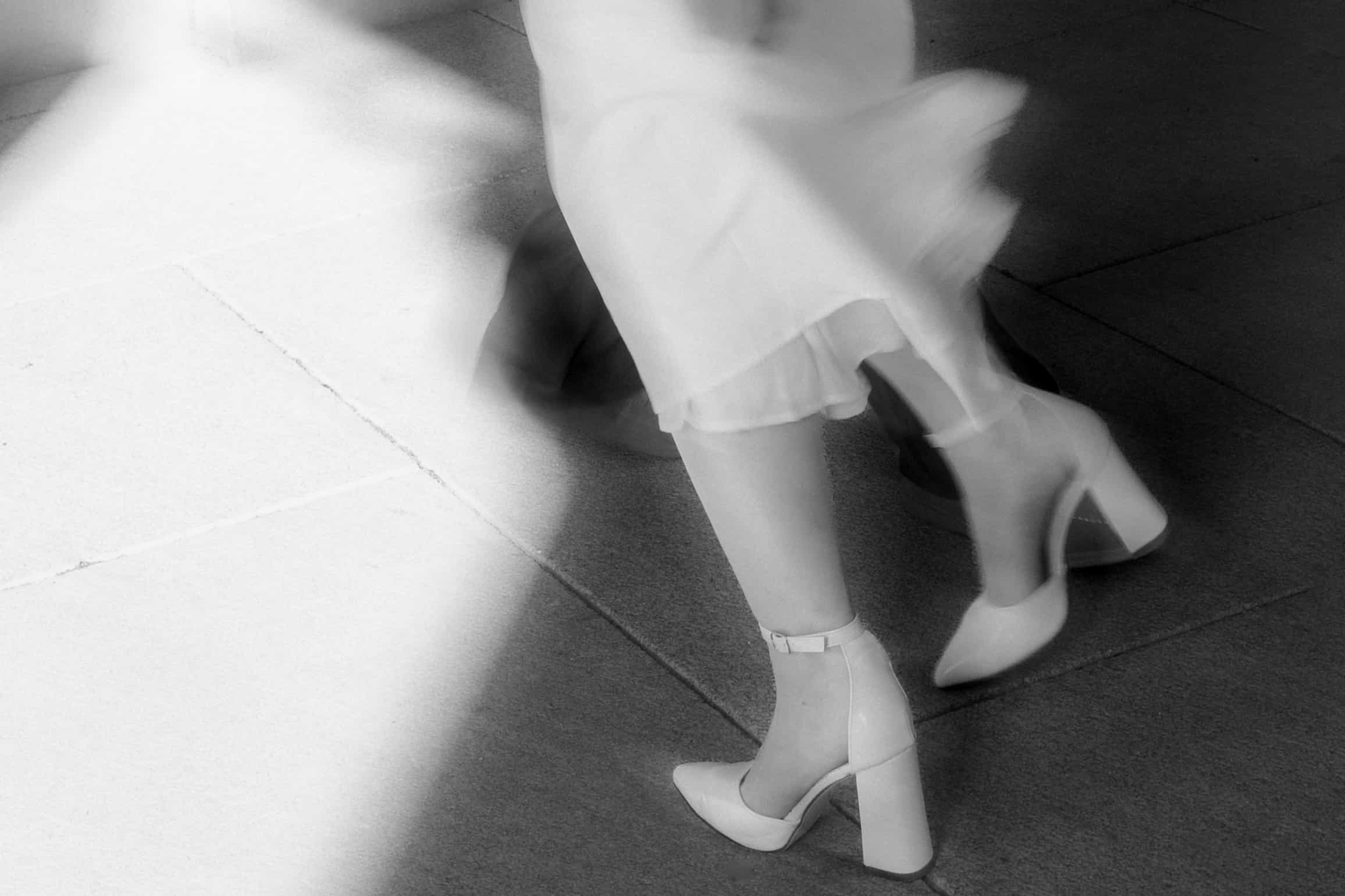 Black-and-white motion-blur photo of a bride's white heels stepping across stone paving, dress hem blurred