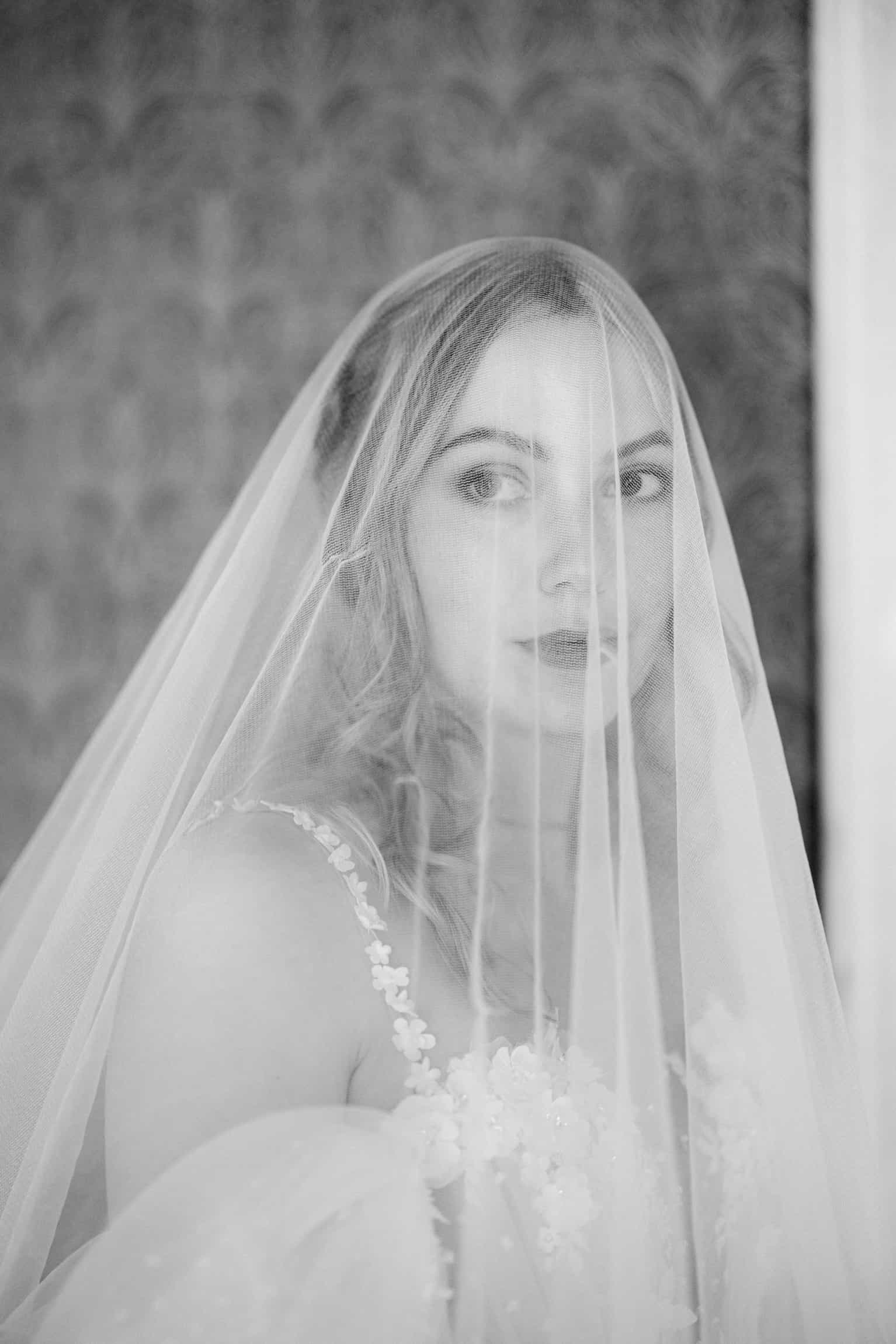 Bride in an intricate lace veil gazing downward, black-and-white portrait against a neutral background