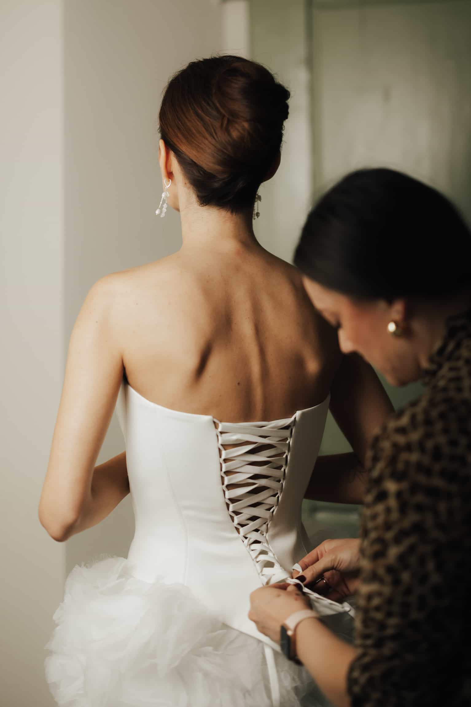 Back view of bride in a white wedding dress being helped with corset lacing during getting-ready