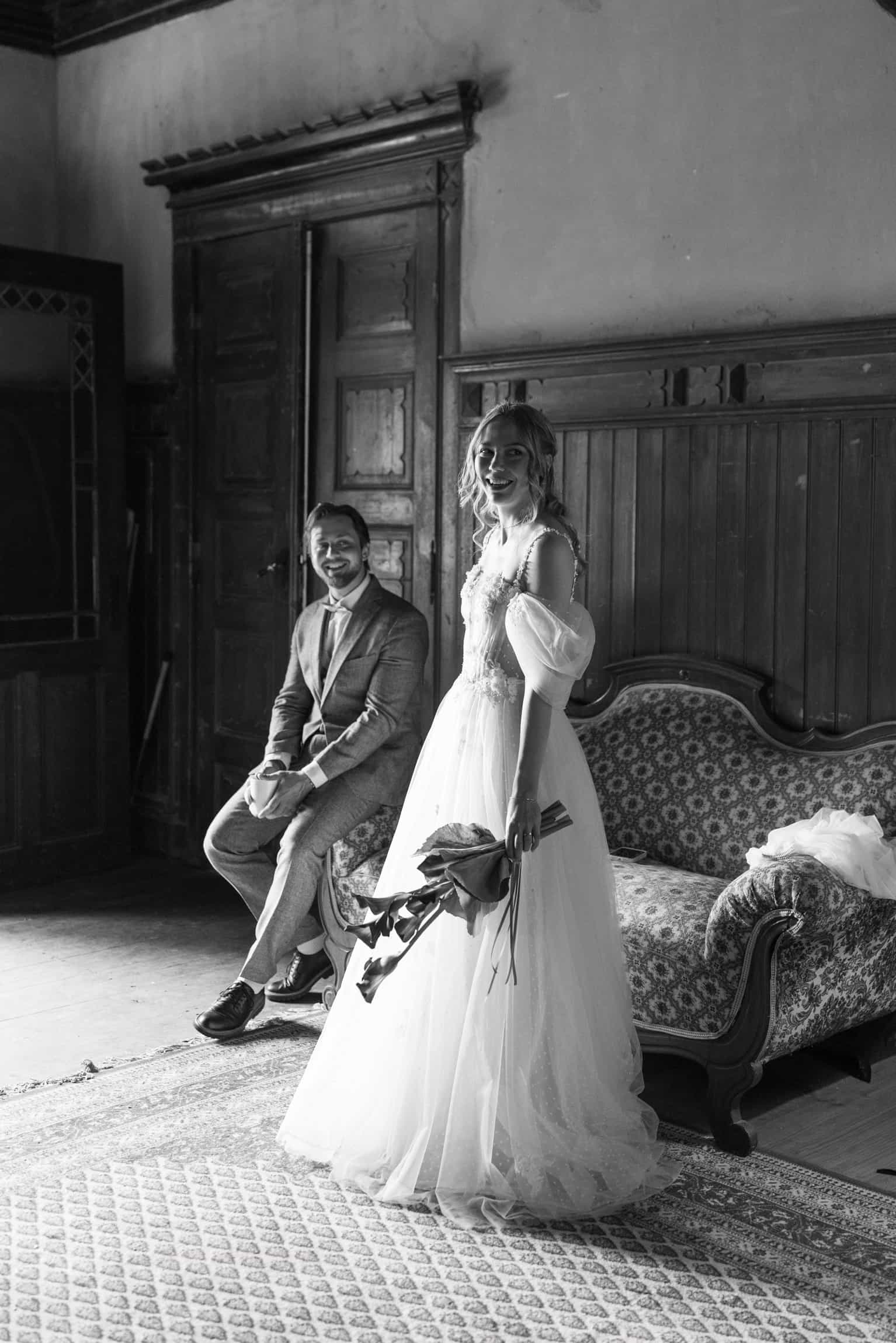 Bride beside groom on a velvet settee in a minimalist vintage room, timeless black-and-white wedding photograph