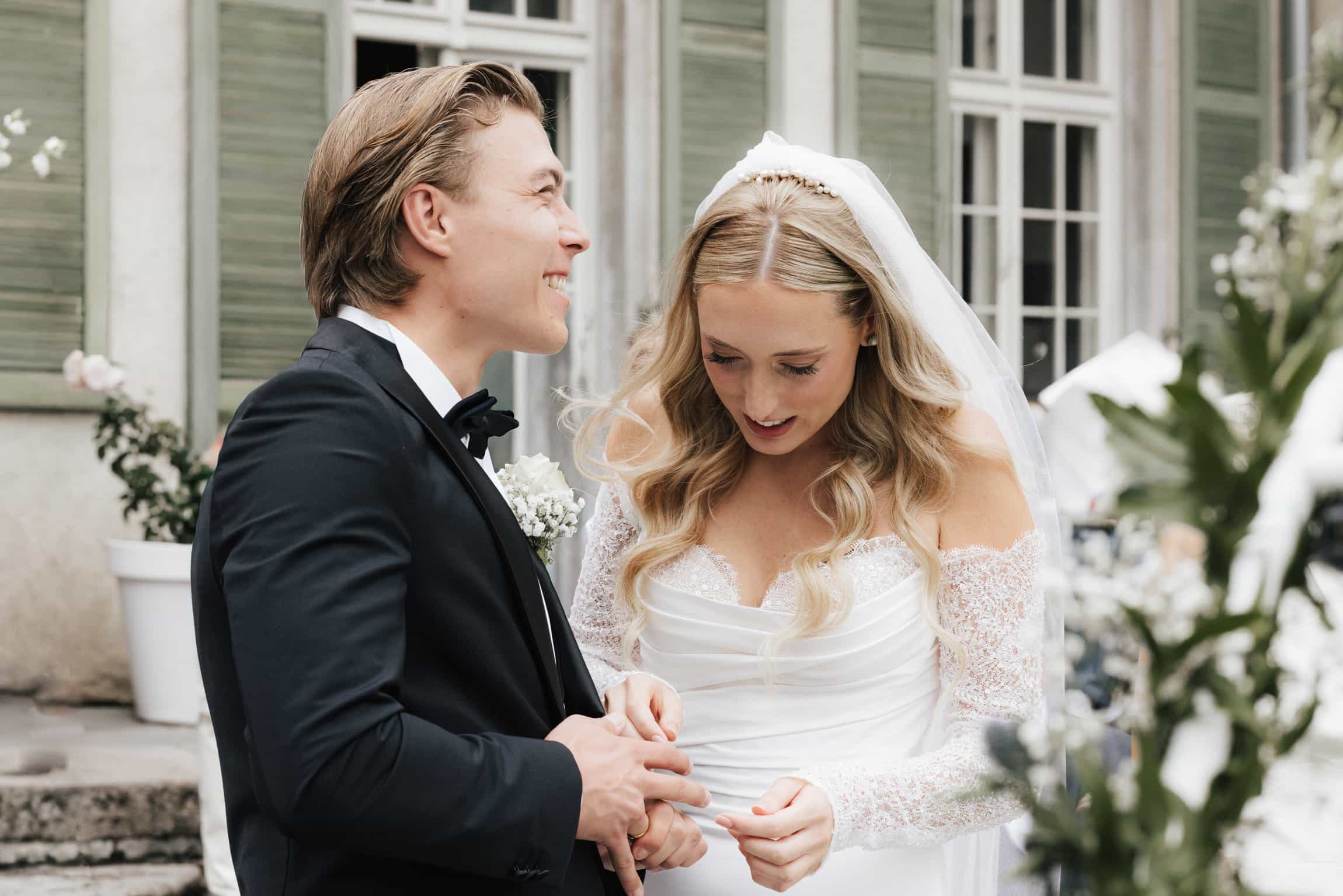 Bride in white lace dress and veil holding hands with groom in a dark suit in an elegant courtyard