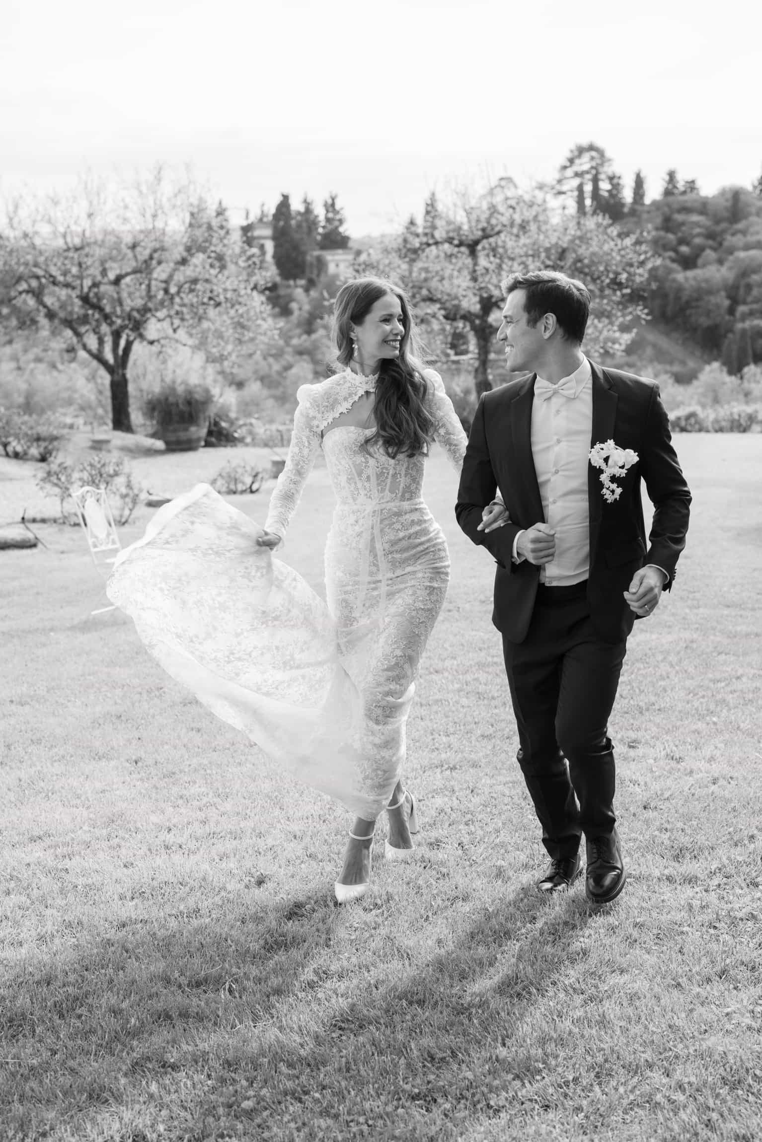 Bride in flowing white dress and veil walking arm in arm with groom through a sunlit garden