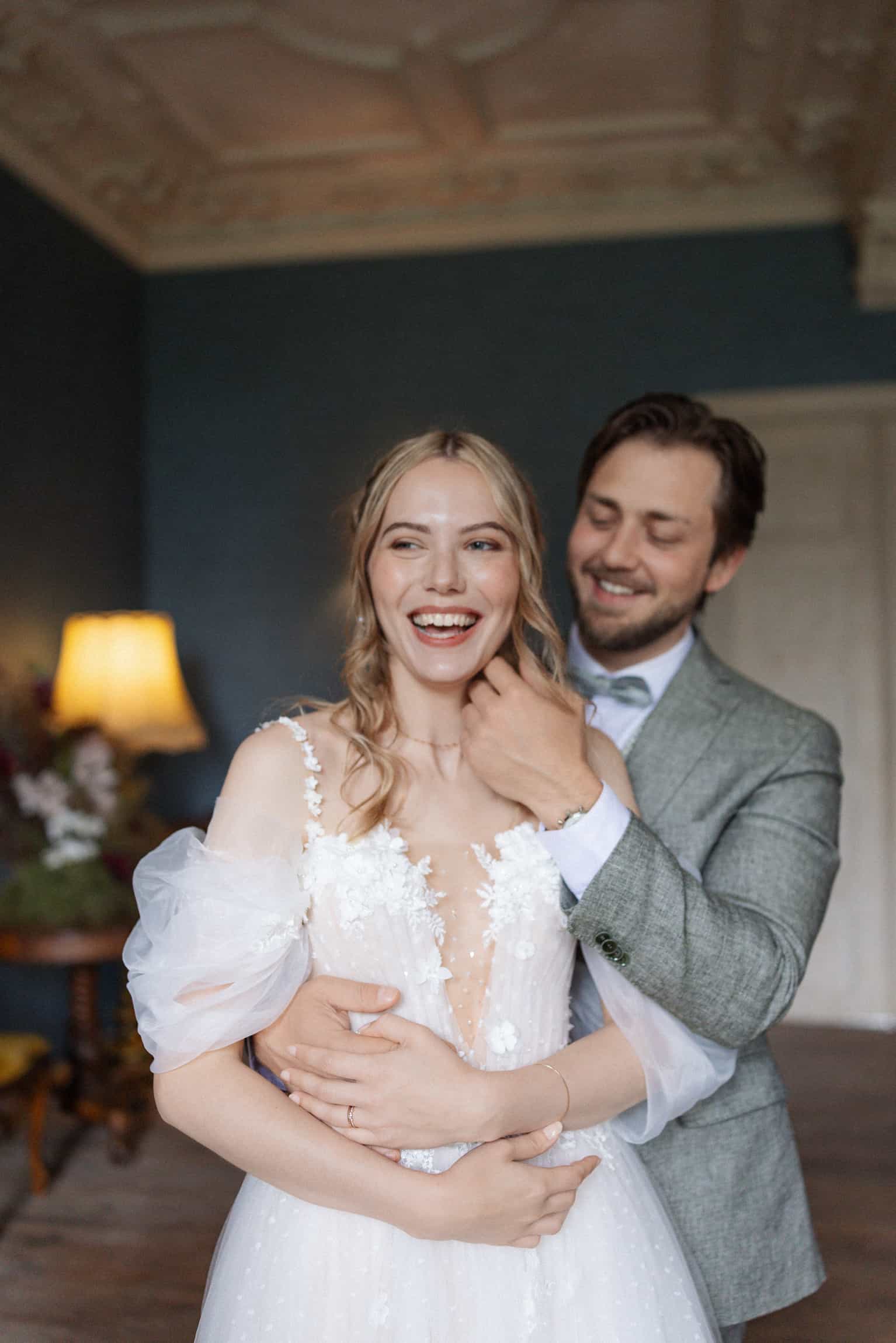 Bride laughing as groom embraces her from behind indoors in warm light, candid wedding moment