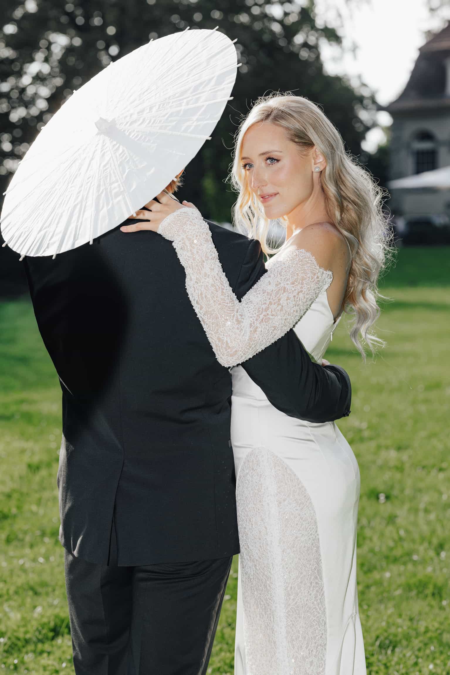 Bride held by groom under a white parasol in evening garden light