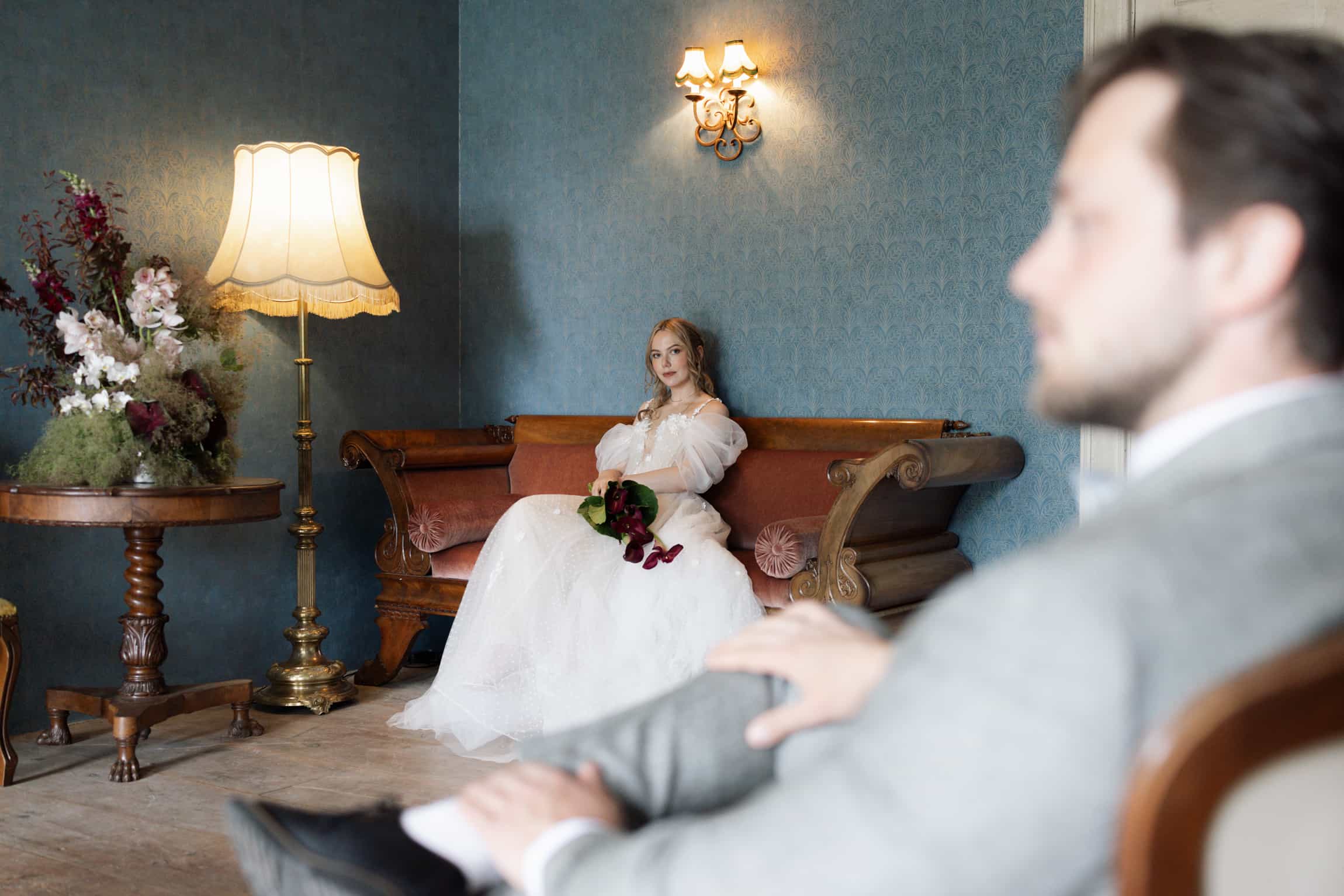 Bride seated on an antique sofa in a blue vintage parlour, viewed past the out-of-focus groom, candid first-look moment