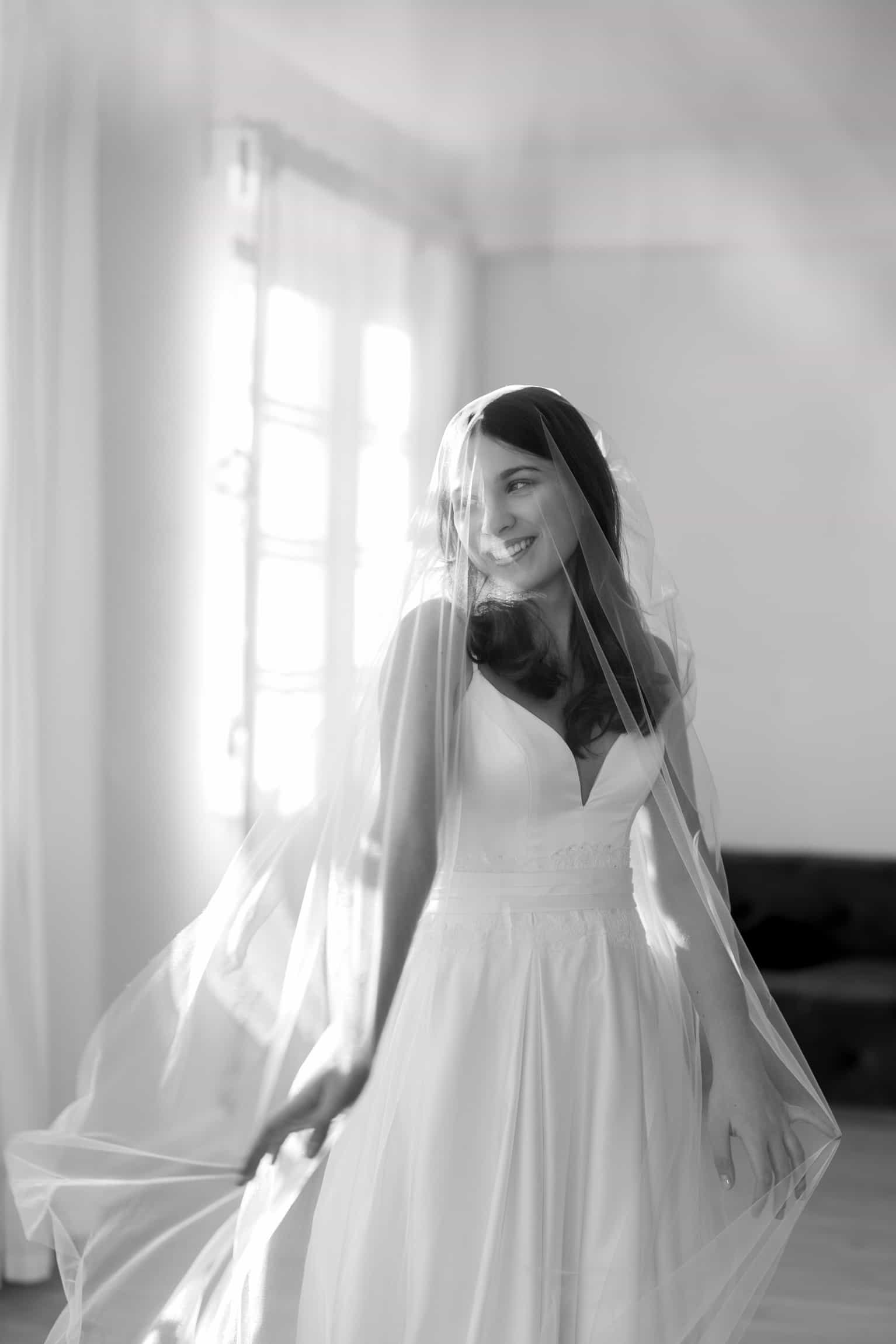 Bride in minimalist white dress and flowing veil, backlit by bright window light in a simple interior