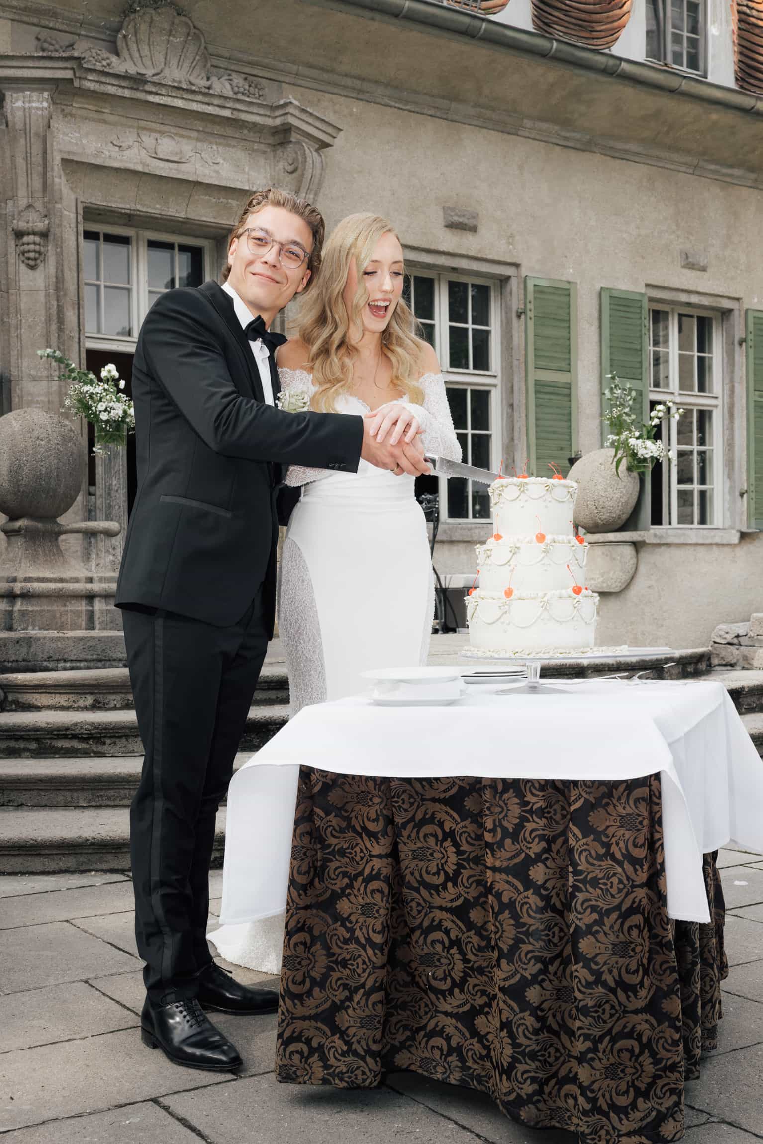 Blonde bride and groom cutting their wedding cake at an outdoor venue with a classical stone building