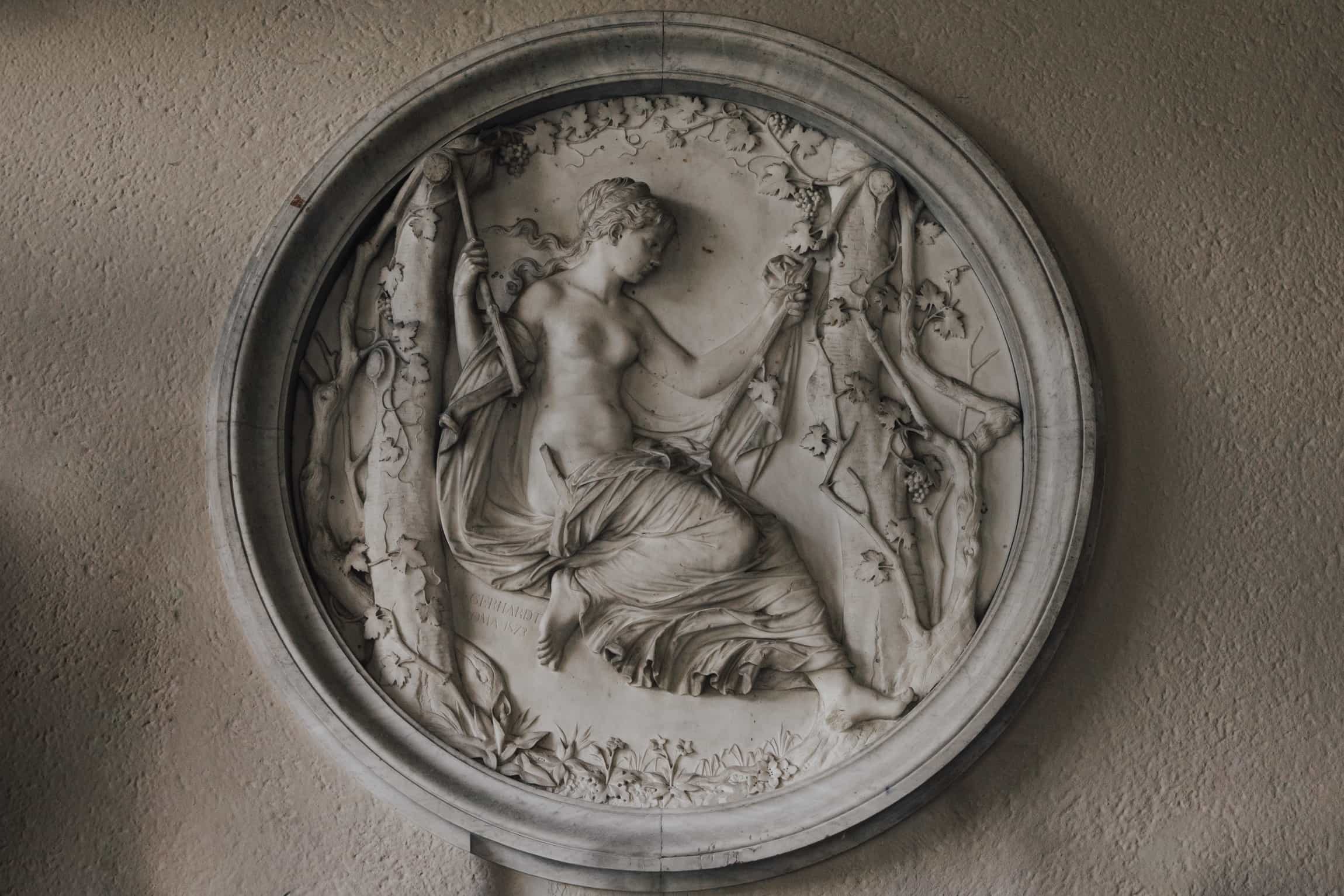 Ornate circular stone medallion with a classical female figure in repose on a neutral wall