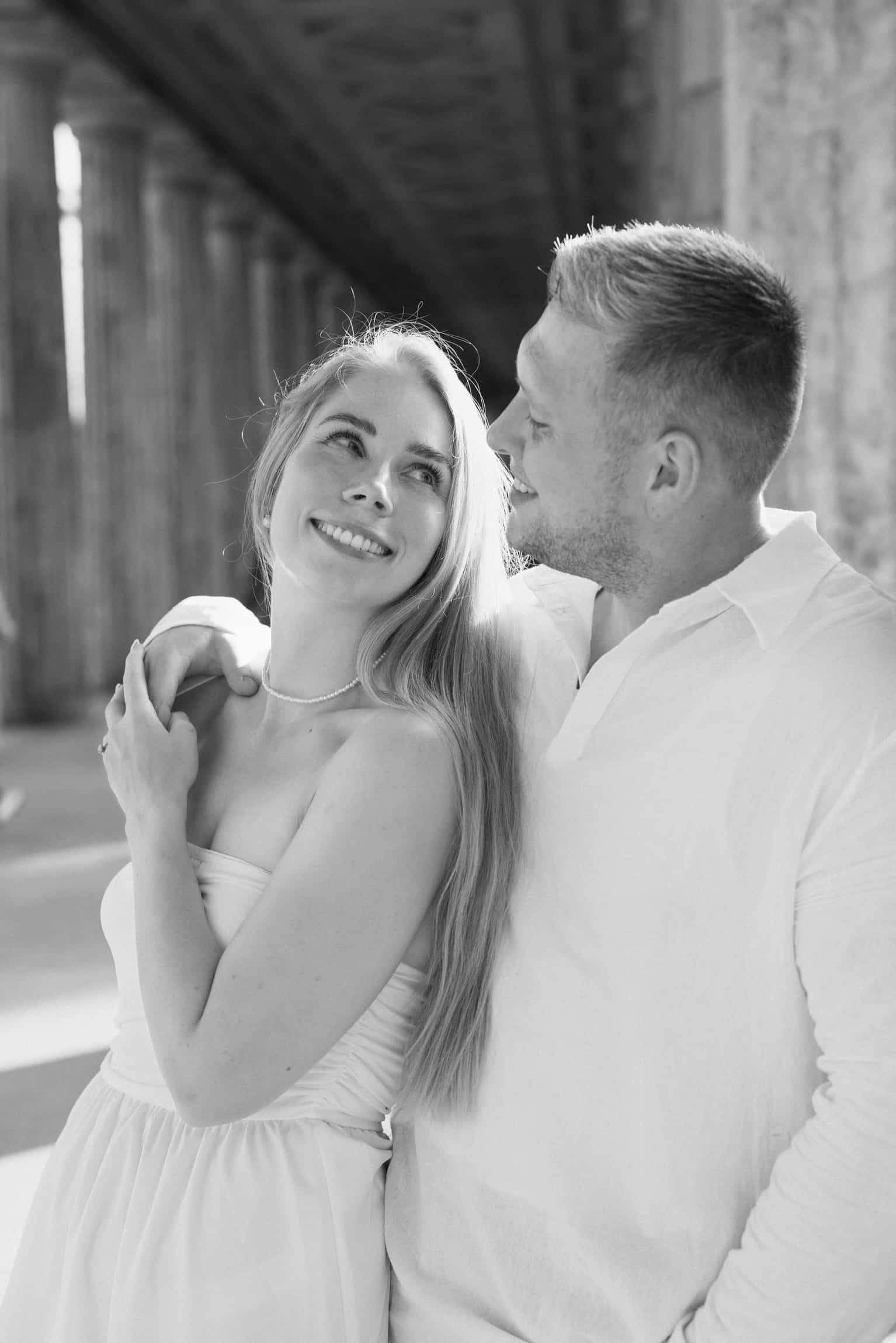 Black-and-white photo of a bride smiling up at her groom as they embrace beneath tall sunlit columns