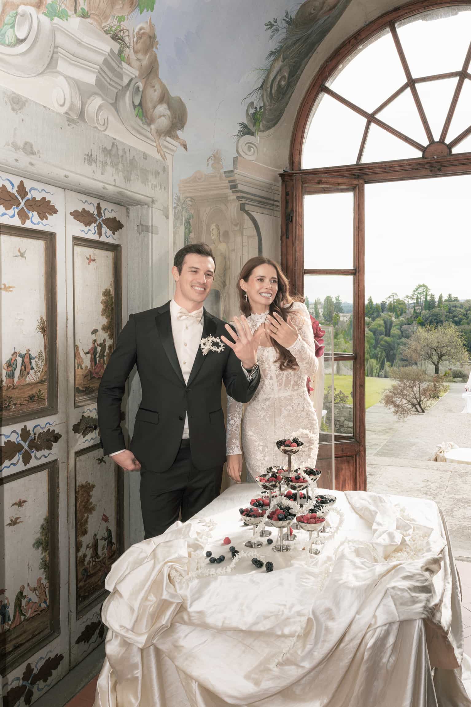 Bride in lace dress and groom at an ornate dessert table with open classical doors framing the view