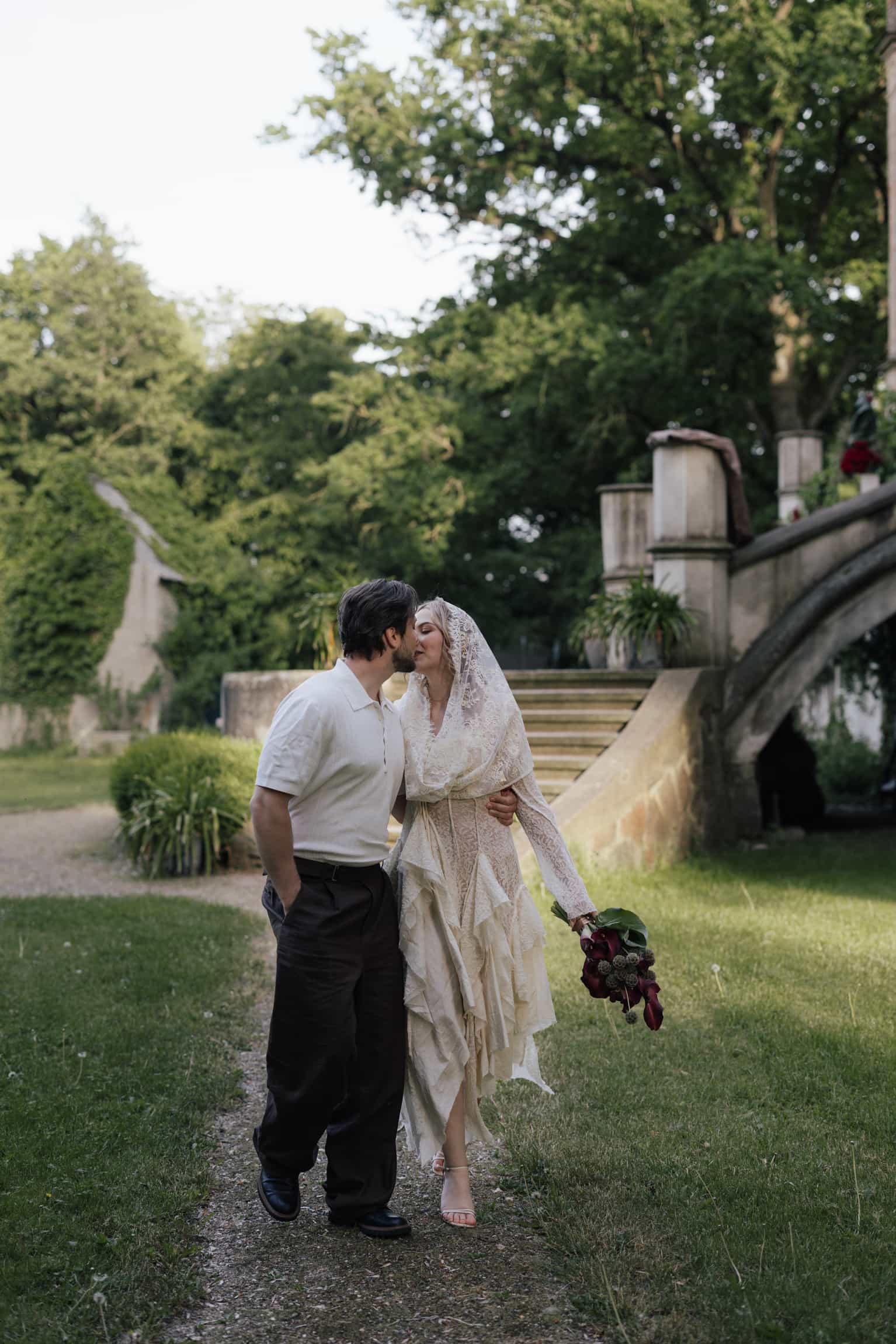 Bride and groom walking together on a gravel path among mature trees and stone structures in a lush garden