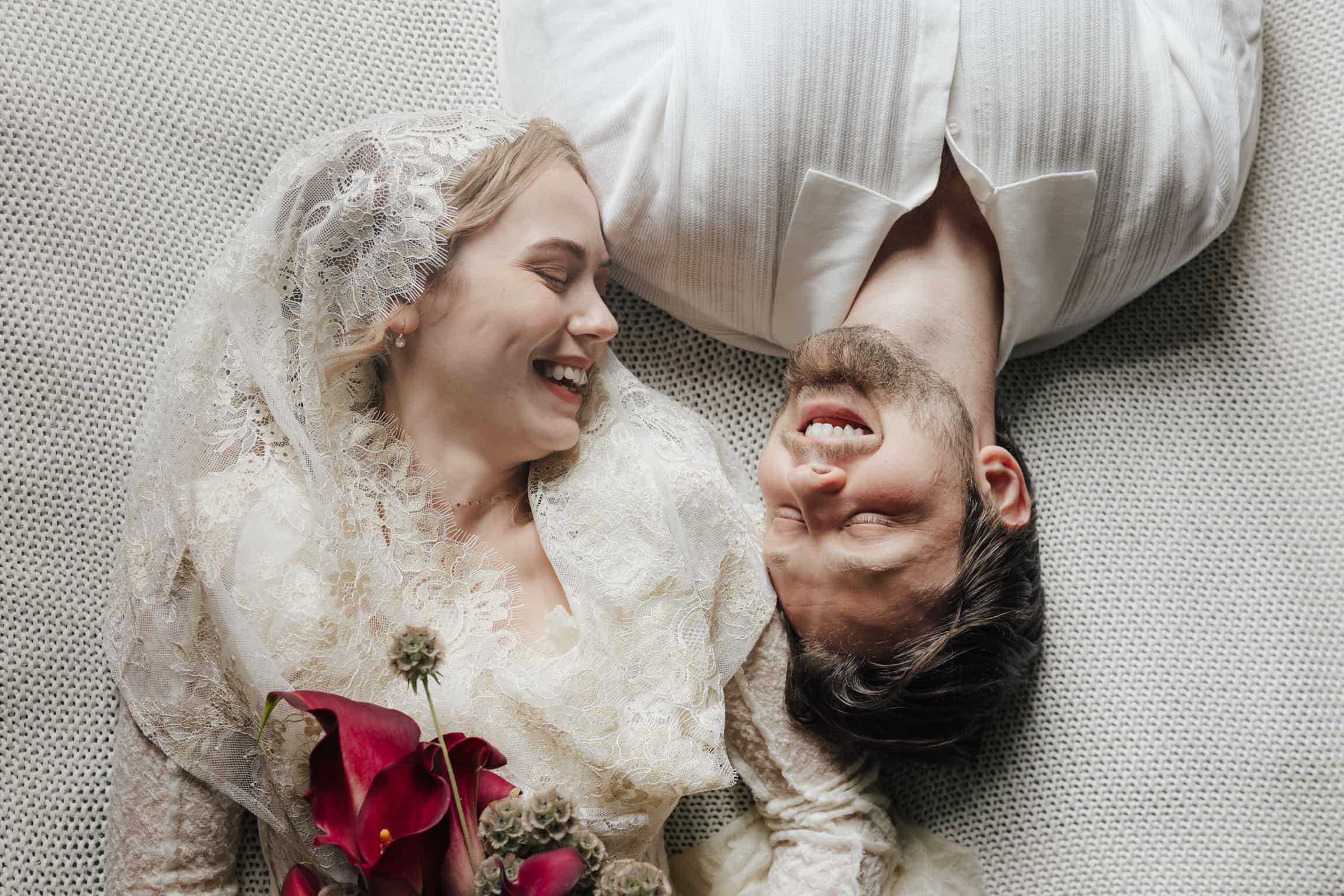Overhead shot of bride and groom lying head to head on white fabric, bride holding red roses, laughing