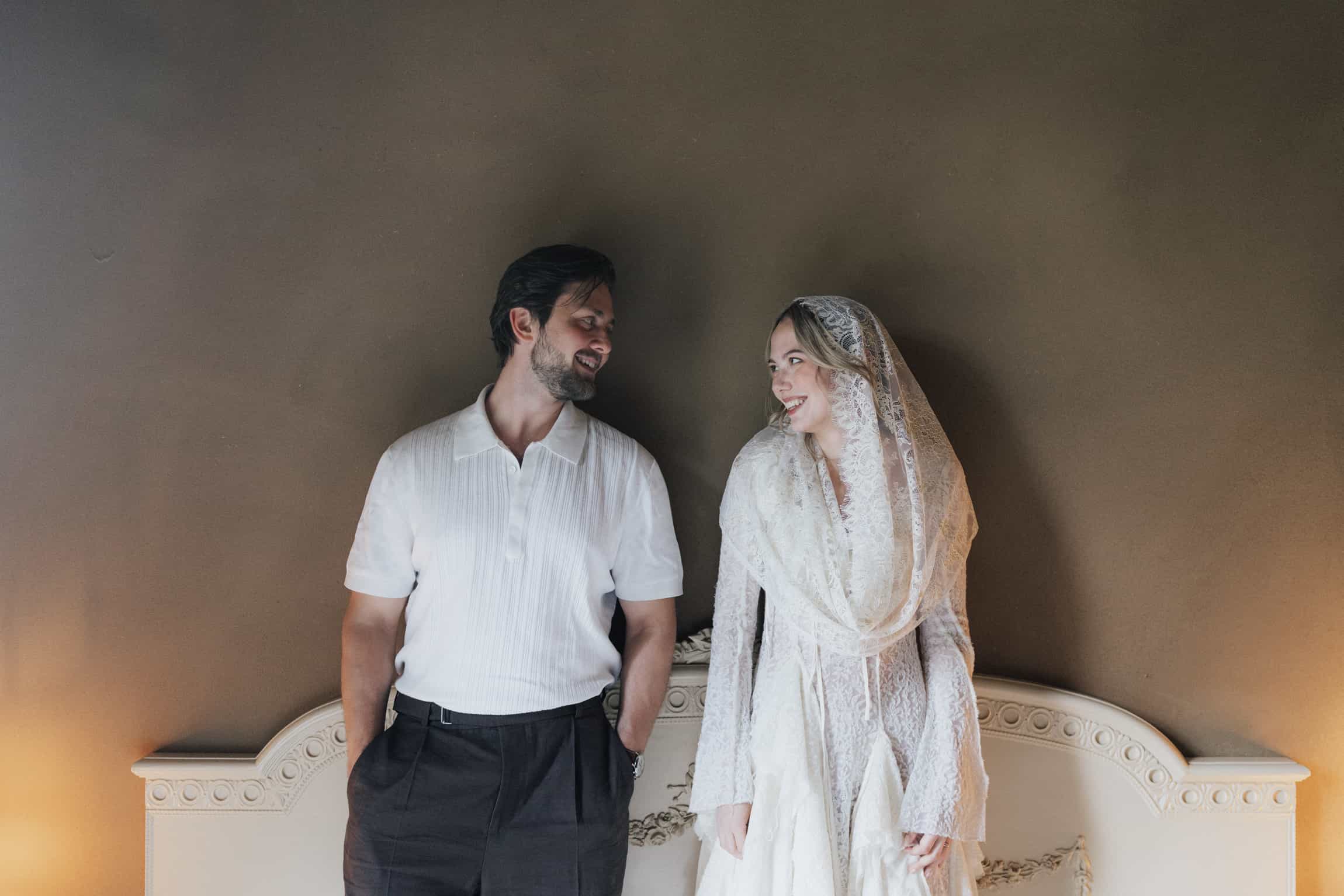 Bride and groom seated together against cream-coloured ornate architecture in soft neutral light