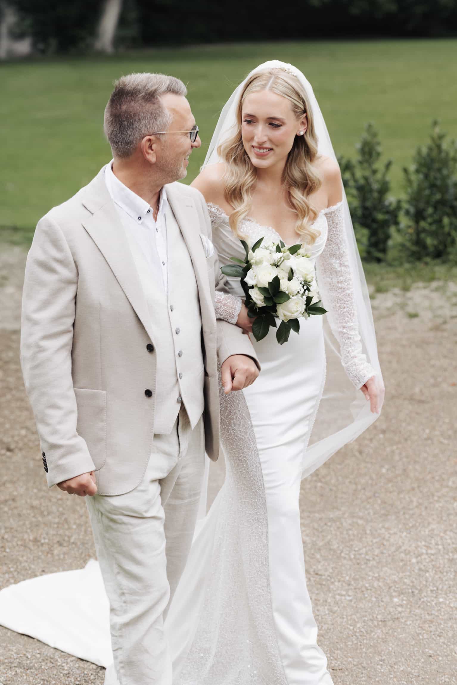 Bride in white dress and groom in cream suit walking a countryside path, natural-light documentary wedding photo