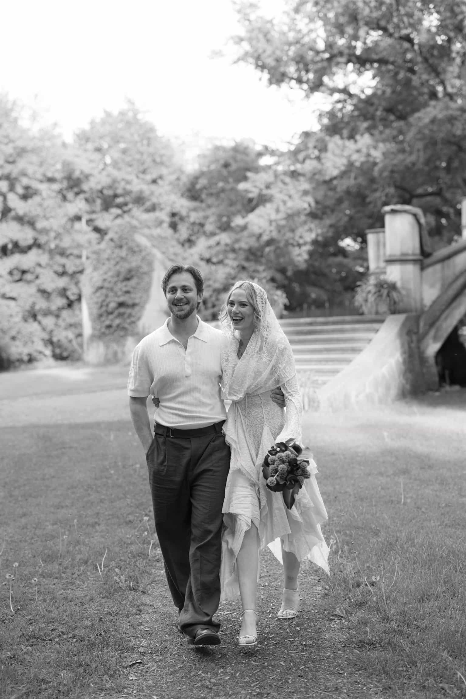 Bride in white dress with veil and groom in light shirt walking together in a leafy garden