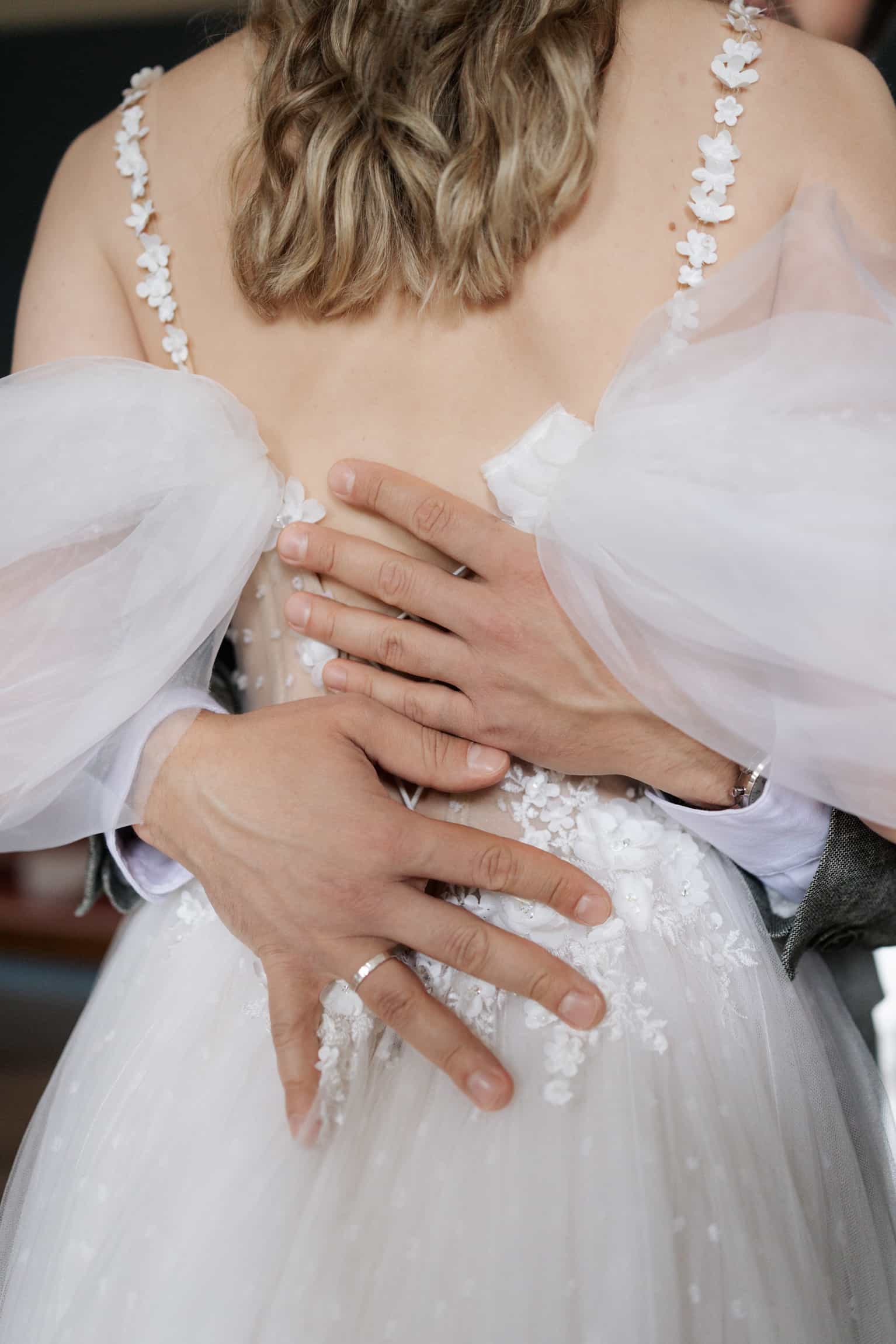 Groom embracing bride from behind, showing the beaded back detail of her white wedding dress