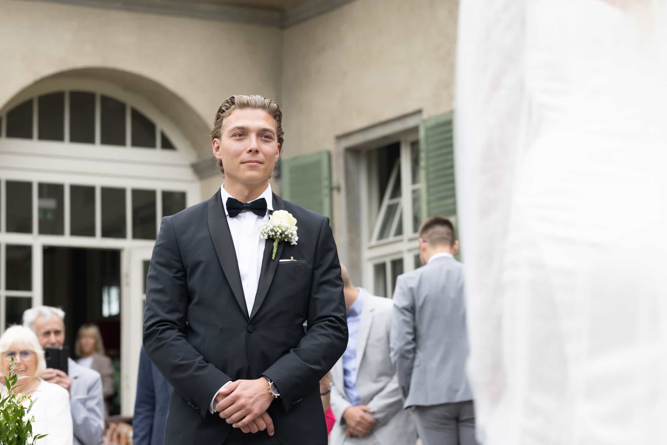 Groom in black tuxedo and bow tie standing alone before arched architectural doorways and columns