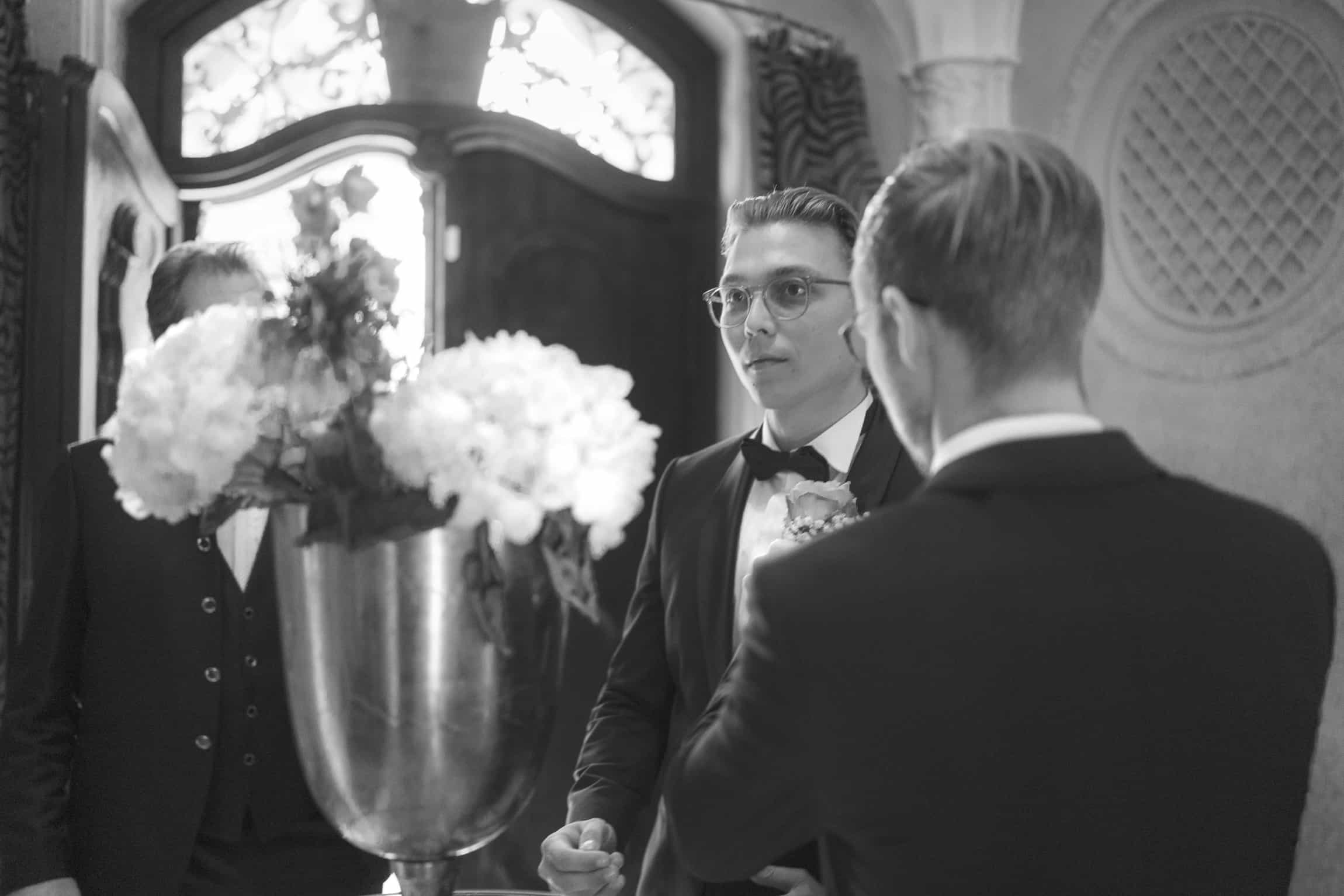 Groom in dark suit and glasses talking with the best man near an ornate doorway with white flowers