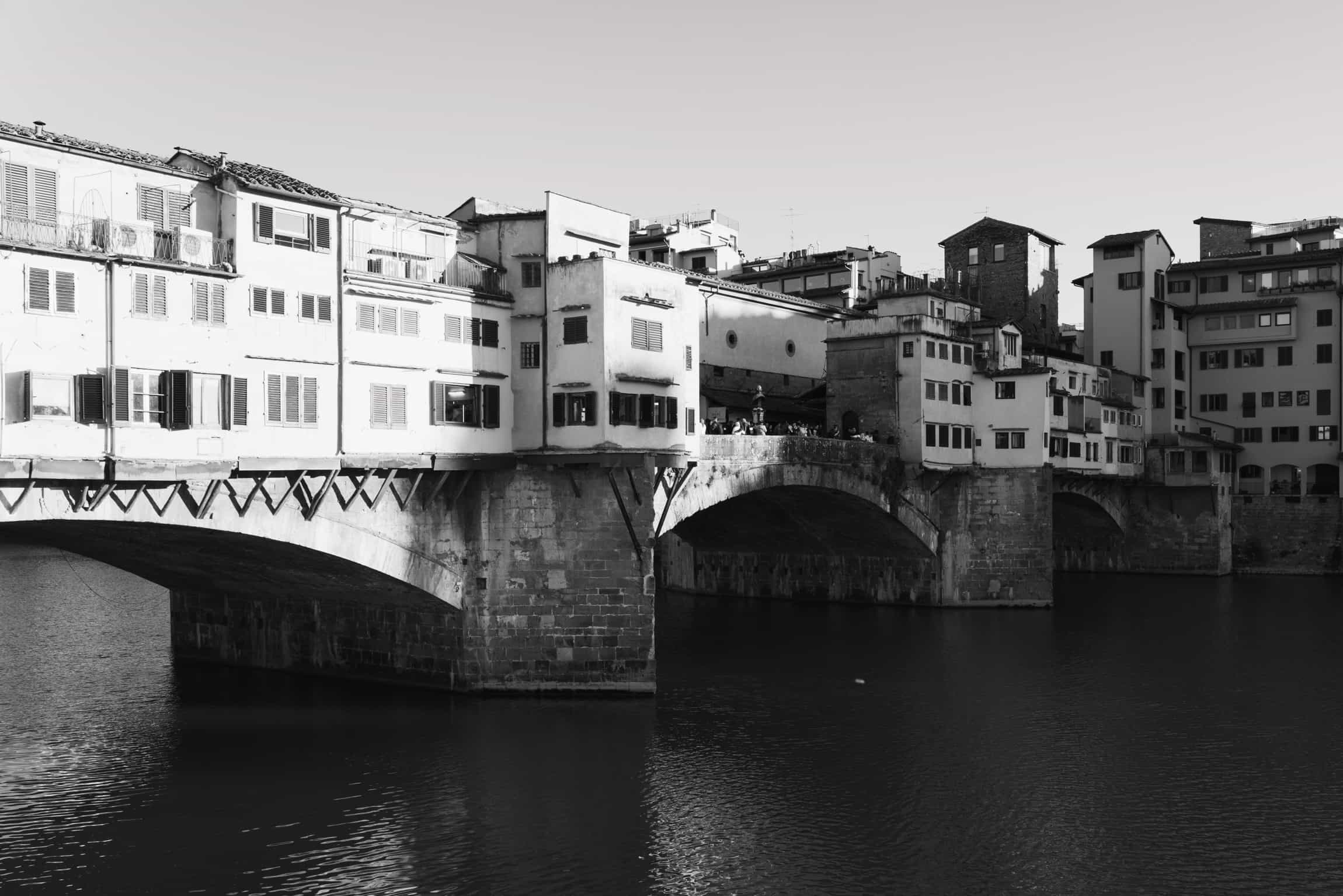 Black-and-white photograph of a timeless arched stone bridge with riverside shops and historic buildings