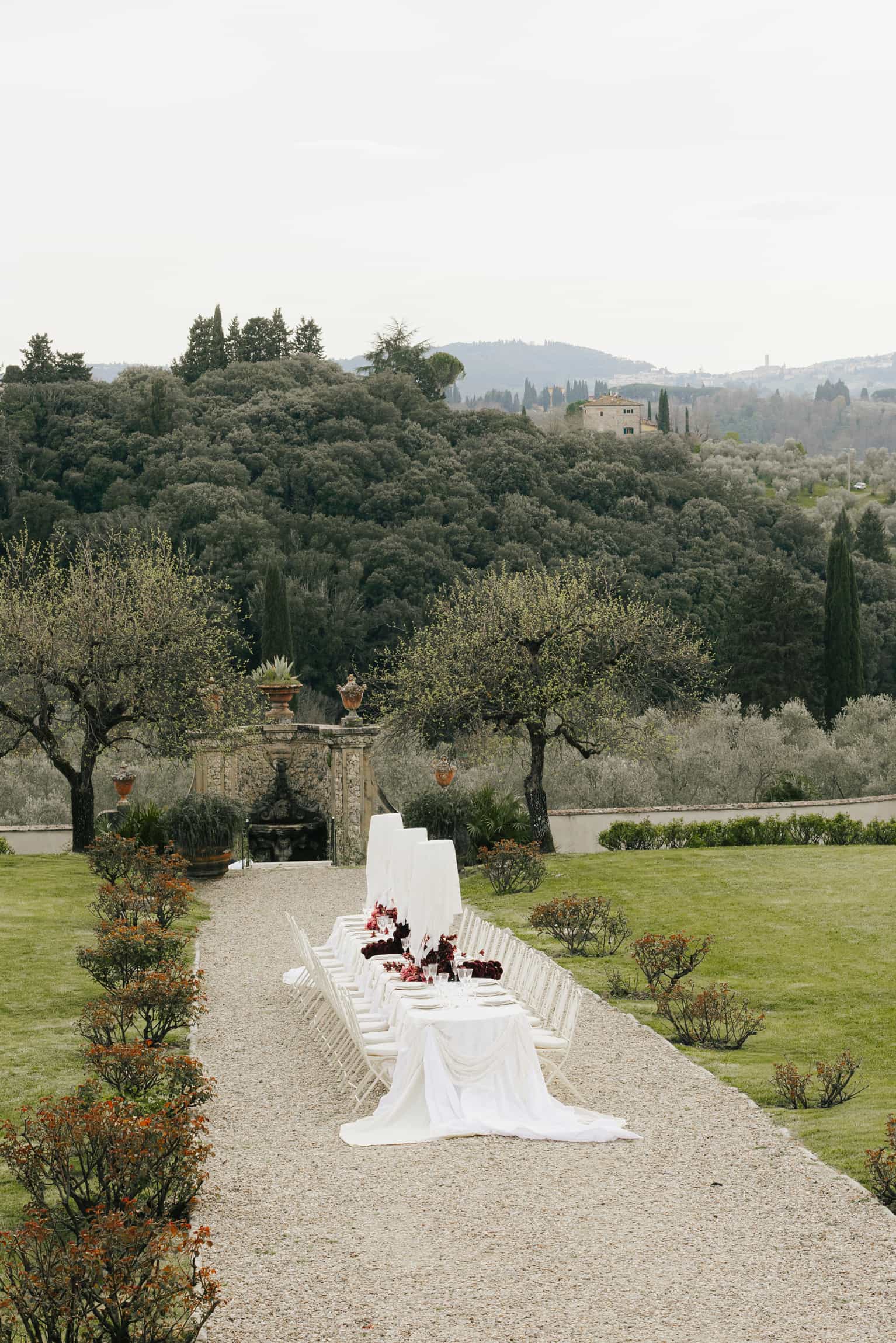 Long minimalist white-draped table in a formal garden with olive and cypress trees, intimate wedding dinner