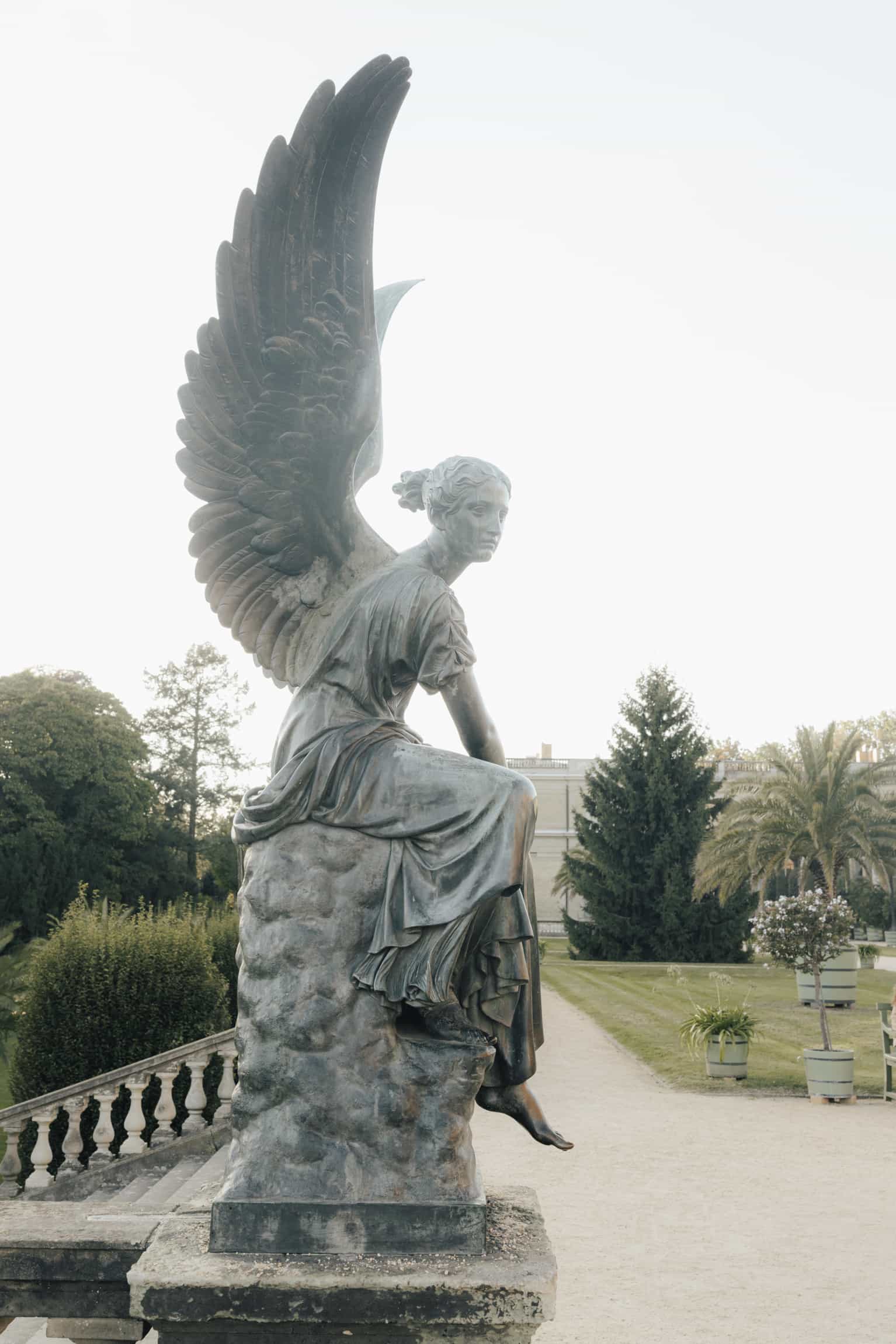 Stone neoclassical angel sculpture with spread wings overlooking formal gardens and distant landscape