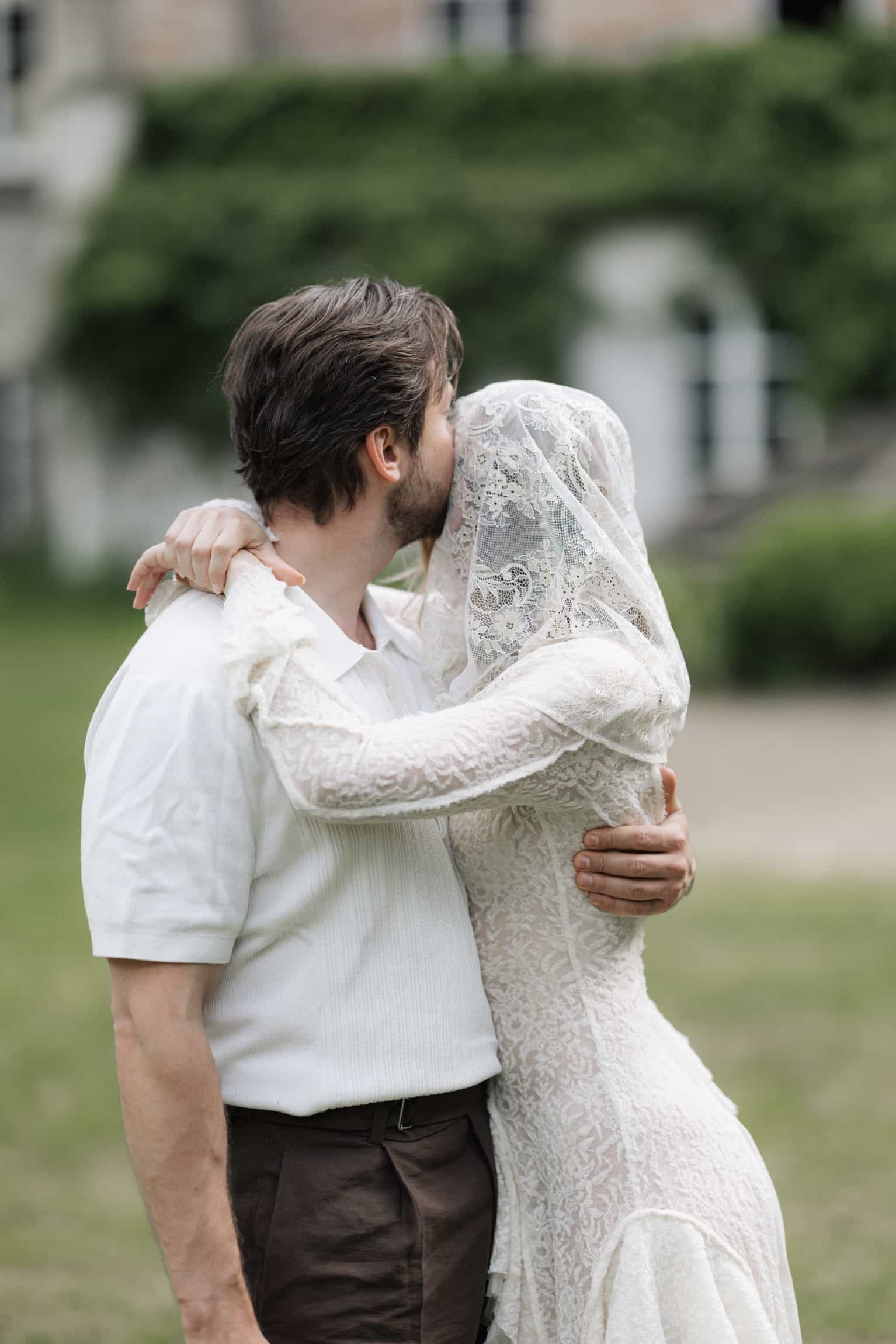 Groom in white shirt embracing bride in lace dress and veil outdoors with green hedges behind