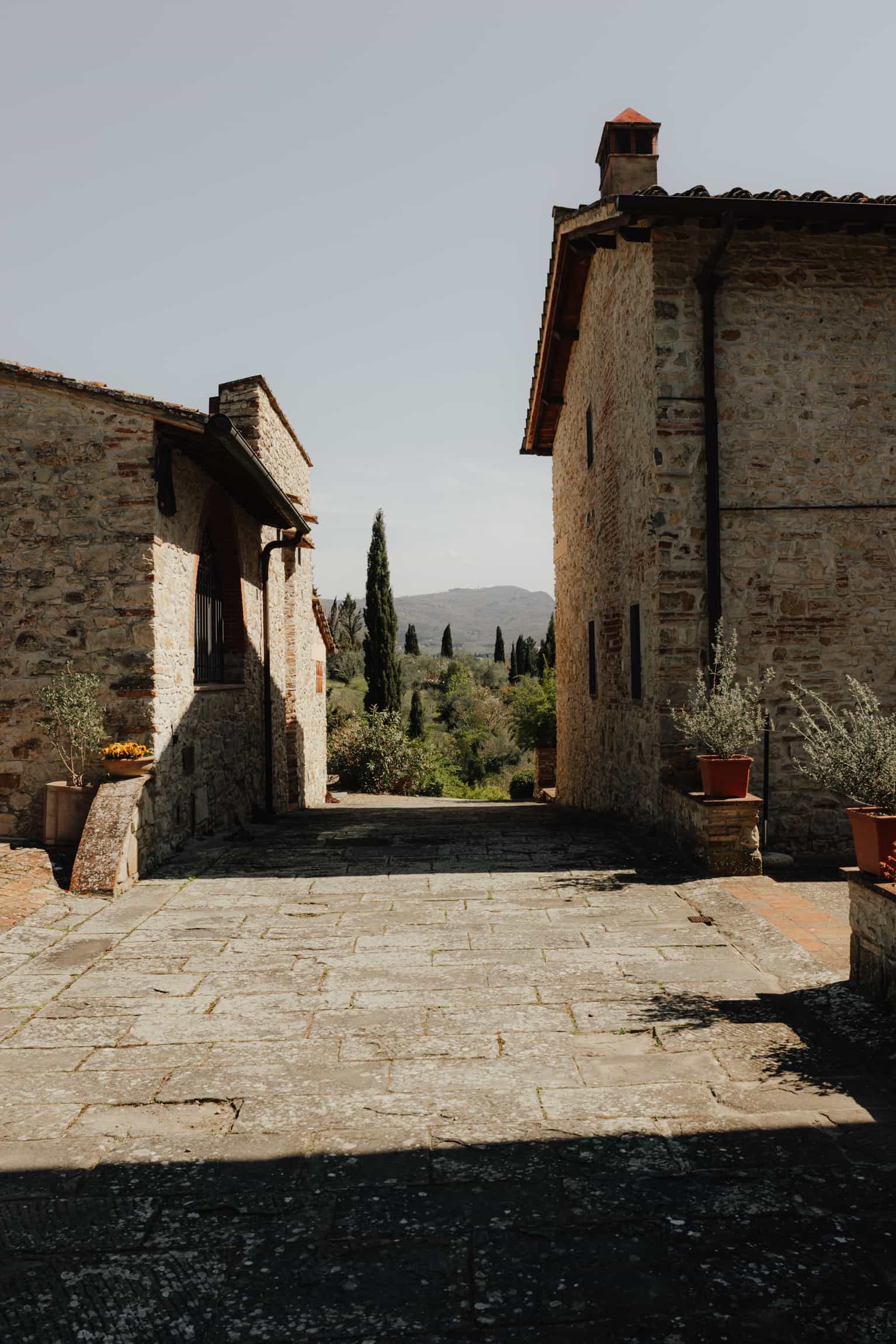 Stone courtyard pathway between rustic buildings with cypress trees in the distance, Tuscan setting