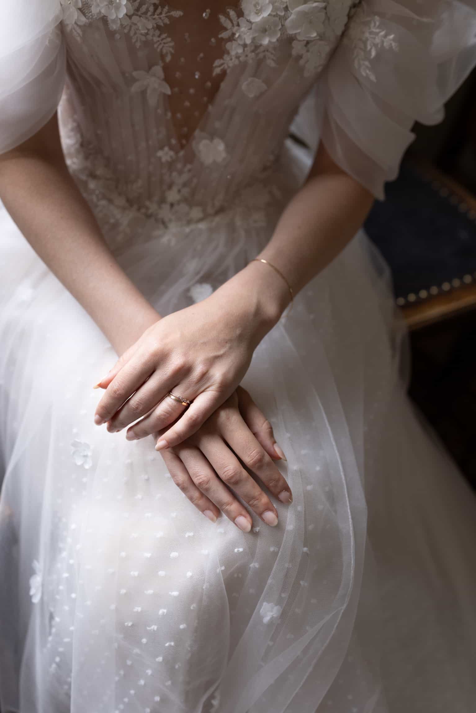 Close-up of bride's hand with wedding ring resting on groom's hand, delicate lace dress above