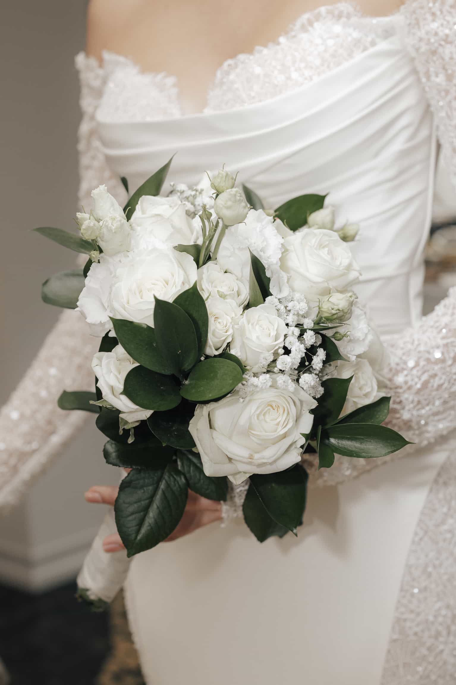 Bride holding a classic white rose bouquet with green foliage, beaded dress detail visible